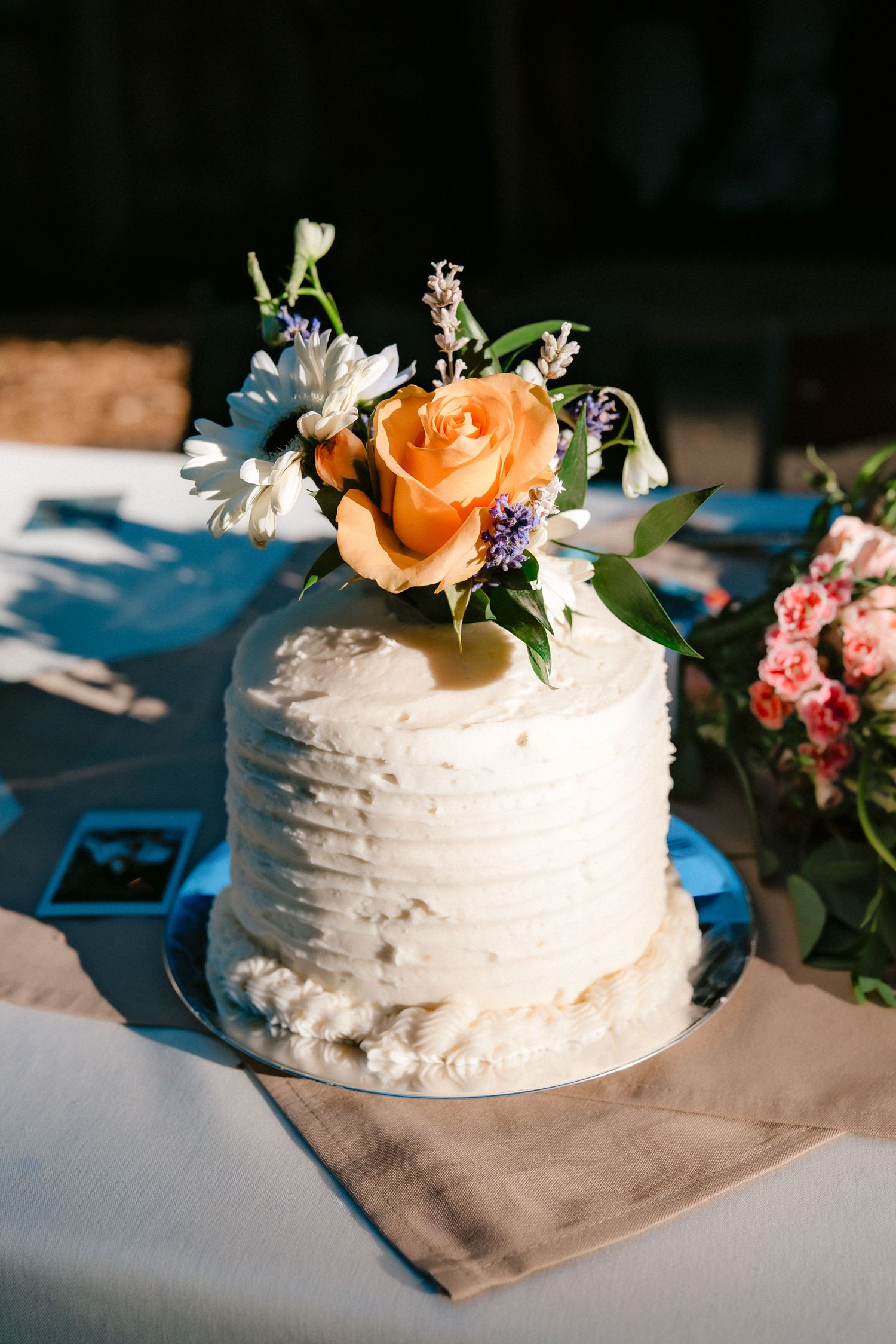 Elegant single-tier white wedding cake topped with an orange rose and wildflowers on a draped table.