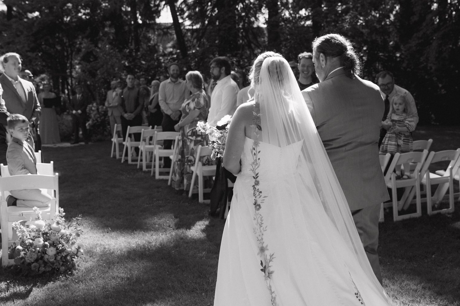 Black and white photo of bride in veil walking down aisle at outdoor garden wedding.