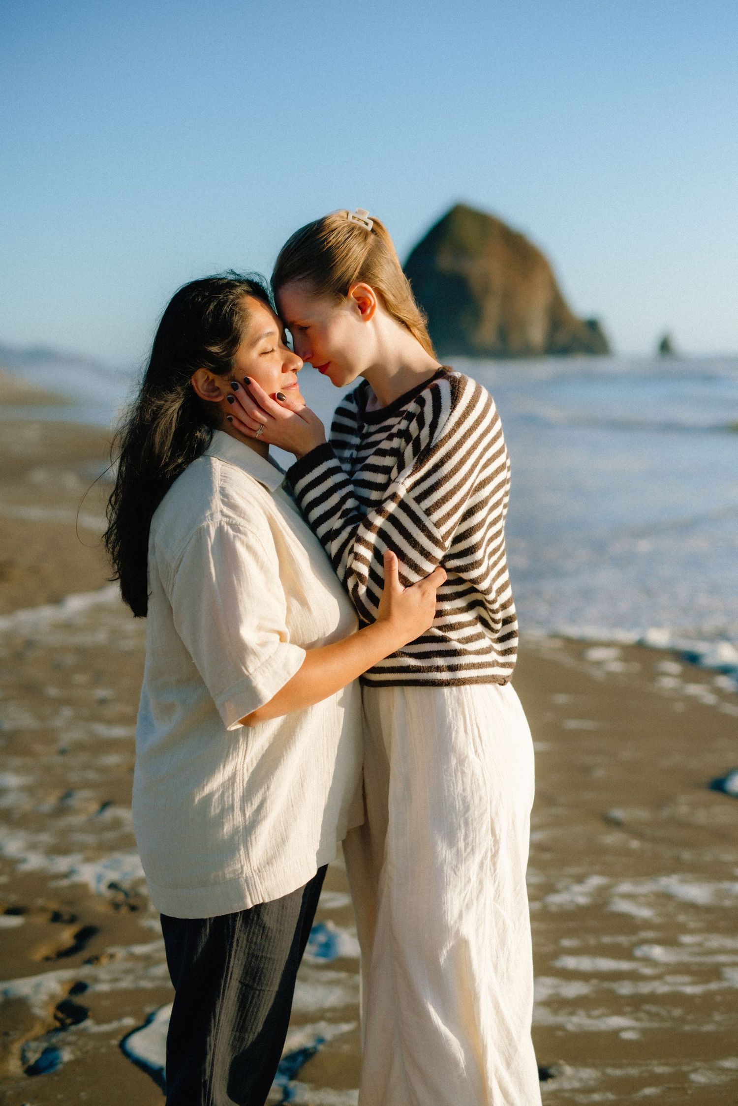 Two women share a romantic moment on a sandy beach with Haystack Rock in the background at golden hour.