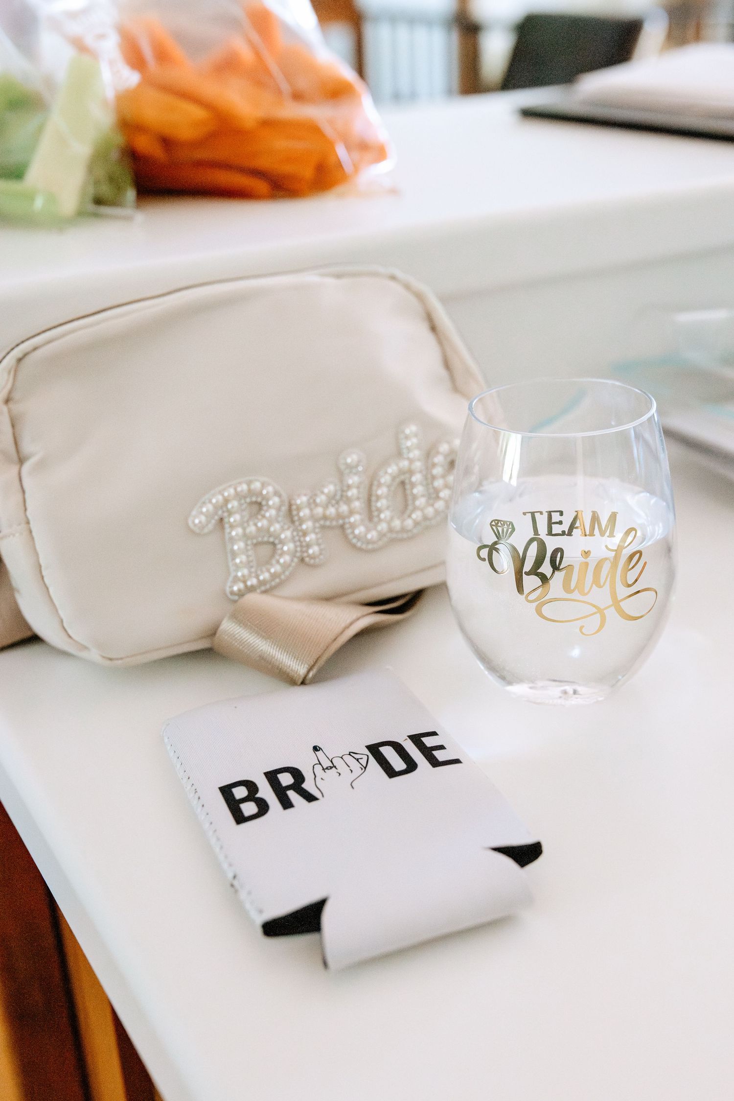 Bridal accessories including a pearl-embroidered cosmetic bag, Team Bride stemless wine glass, and white can koozie on a white surface.