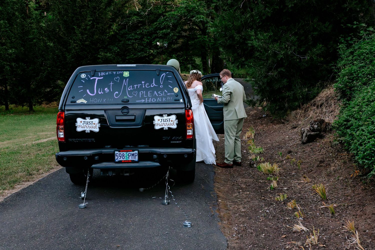 Newlyweds standing beside their Just Married decorated black truck on a wooded driveway after wedding.