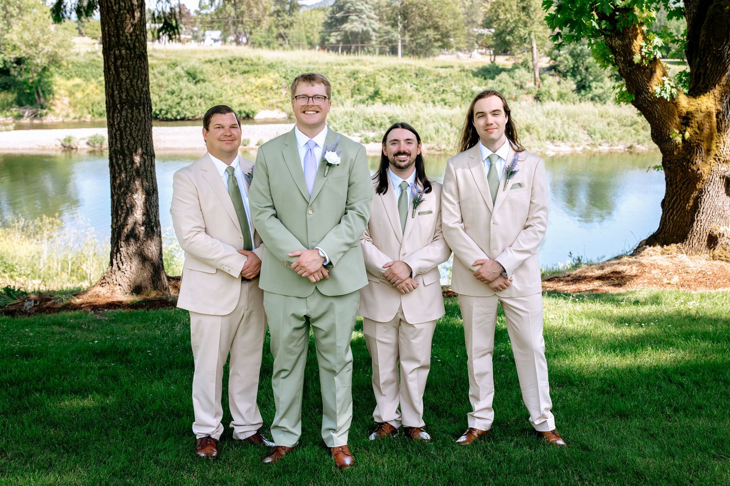 Groom in sage green suit poses with three groomsmen in tan suits beside a lake at an outdoor wedding.