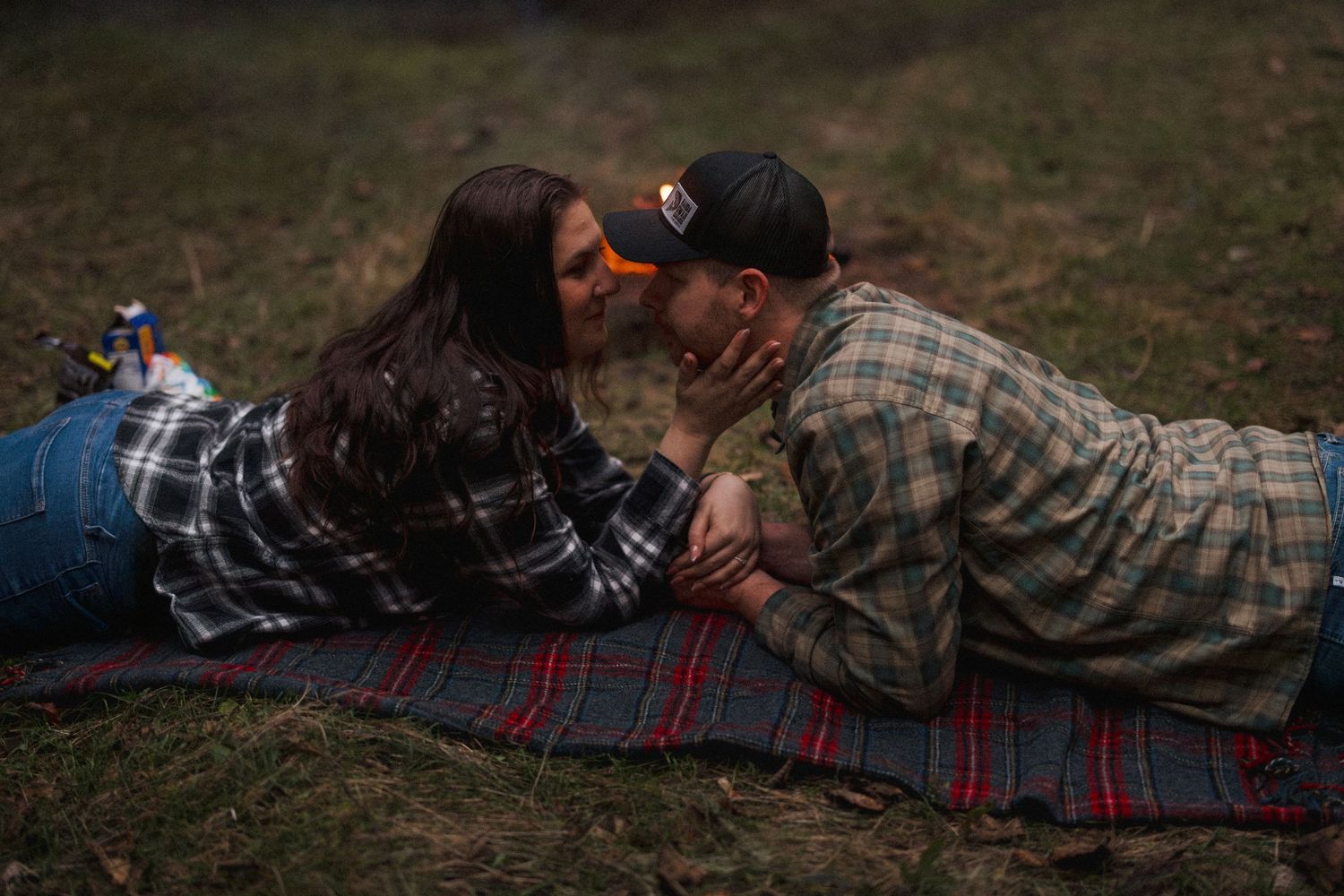 A couple lying on a striped blanket outdoors sharing an intimate moment while wearing plaid shirts.