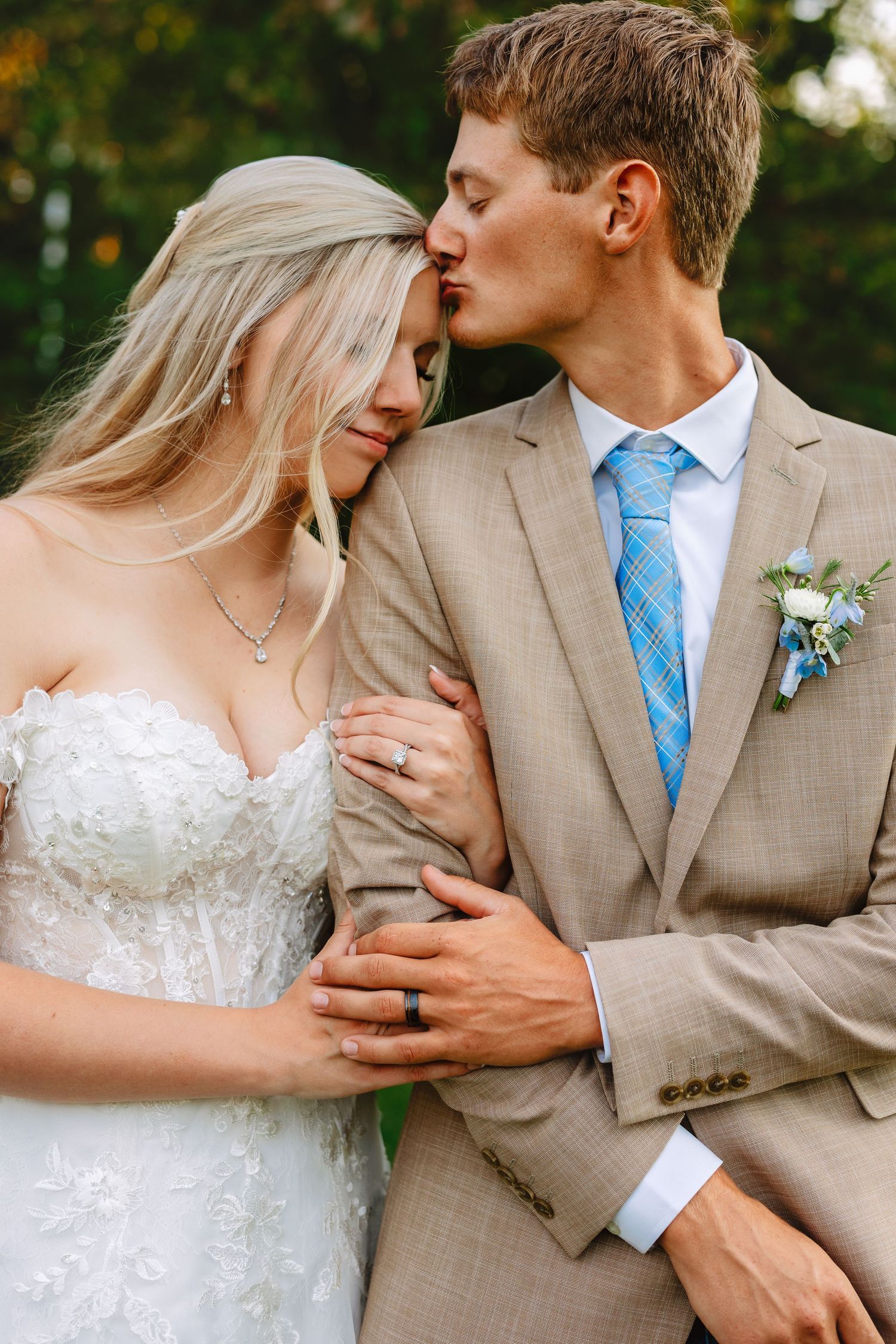 A tender wedding moment outdoors with the couple in a white dress and tan suit with blue tie.