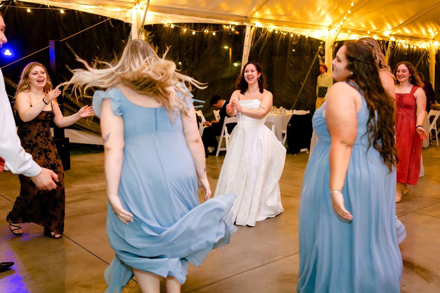 People dancing at a wedding reception under a tent with string lights, featuring bridesmaids in blue dresses.