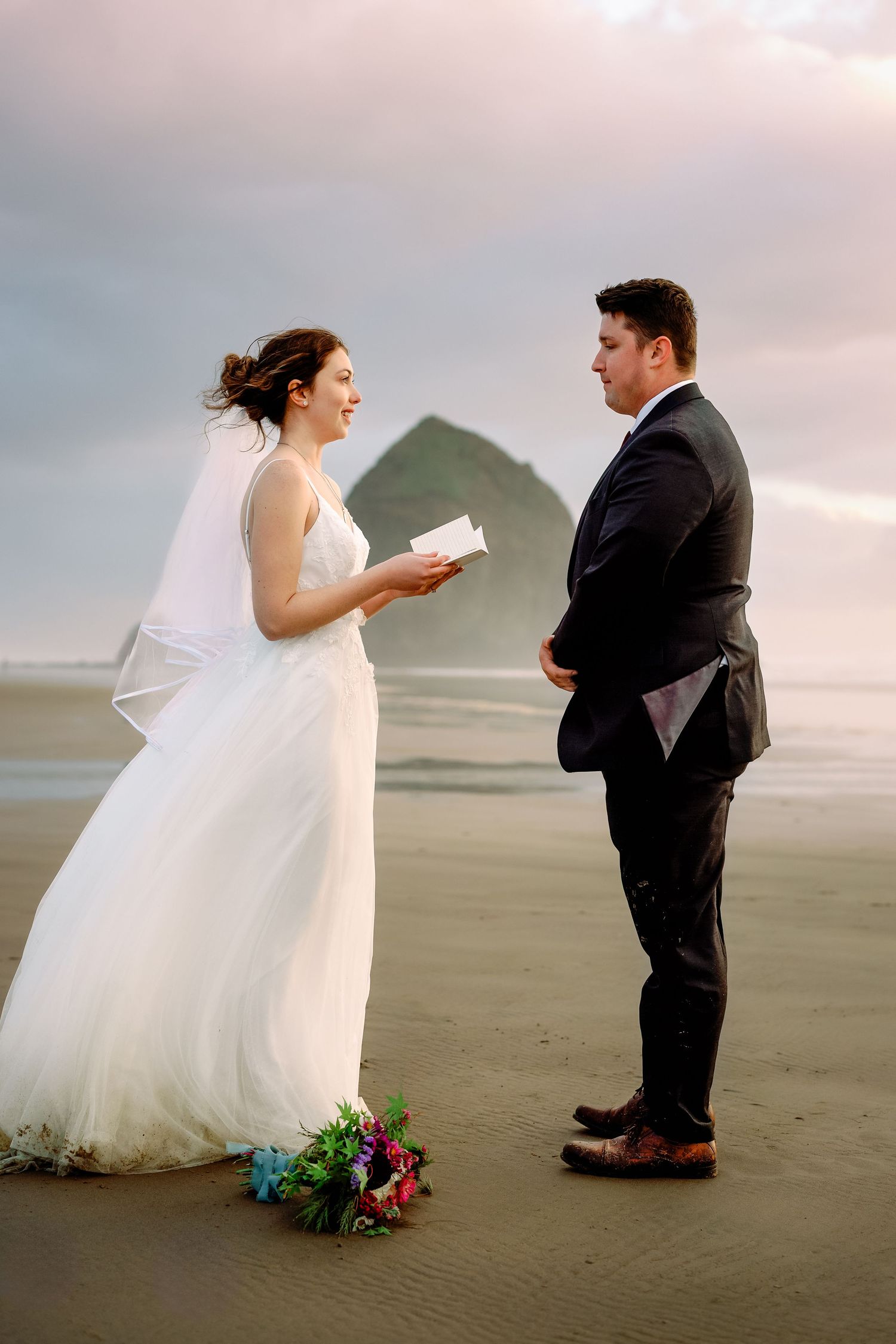 A couple in wedding attire shares a playful moment on a misty beach at sunset.