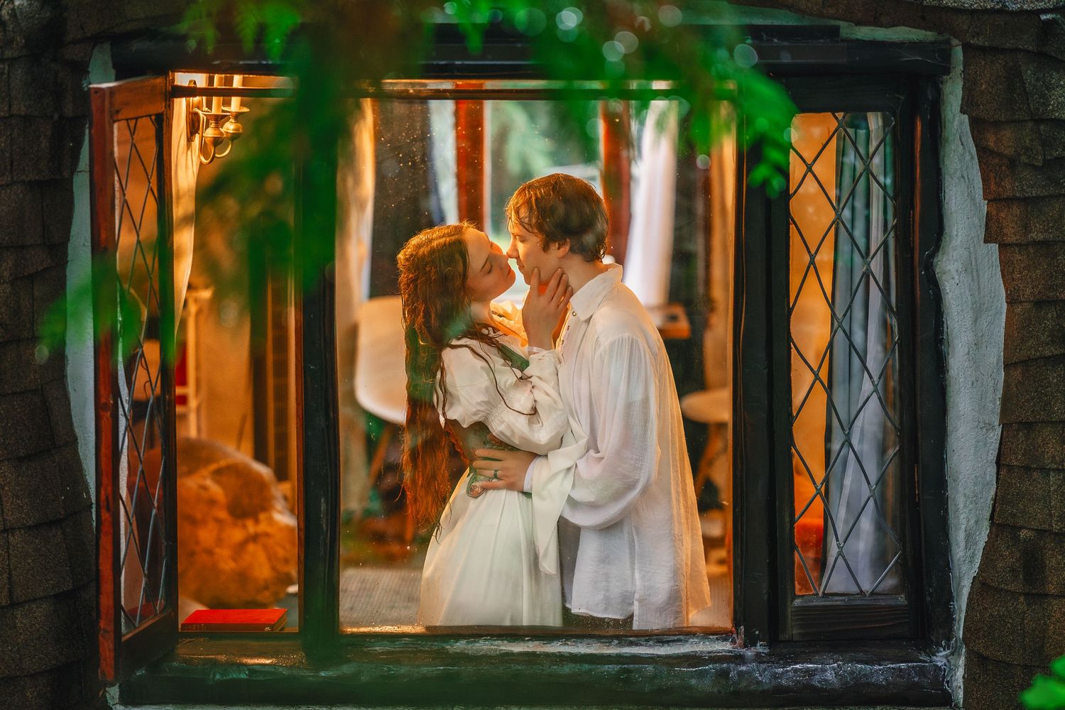 A romantic couple in white clothing share an intimate moment through a window frame with warm lighting and green foliage.