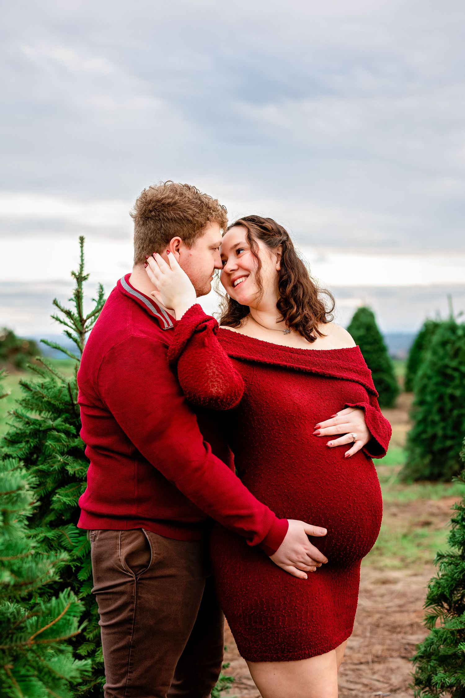 A couple wearing matching red sweaters embrace lovingly among evergreen trees at a Christmas tree farm.