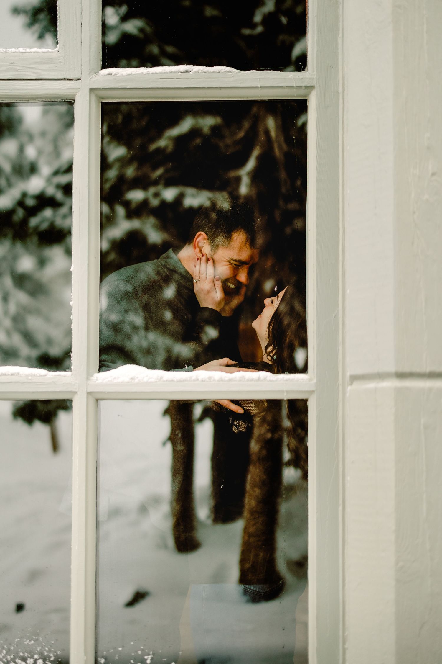 Two people share an intimate moment by a window with snowy evergreen trees visible outside.