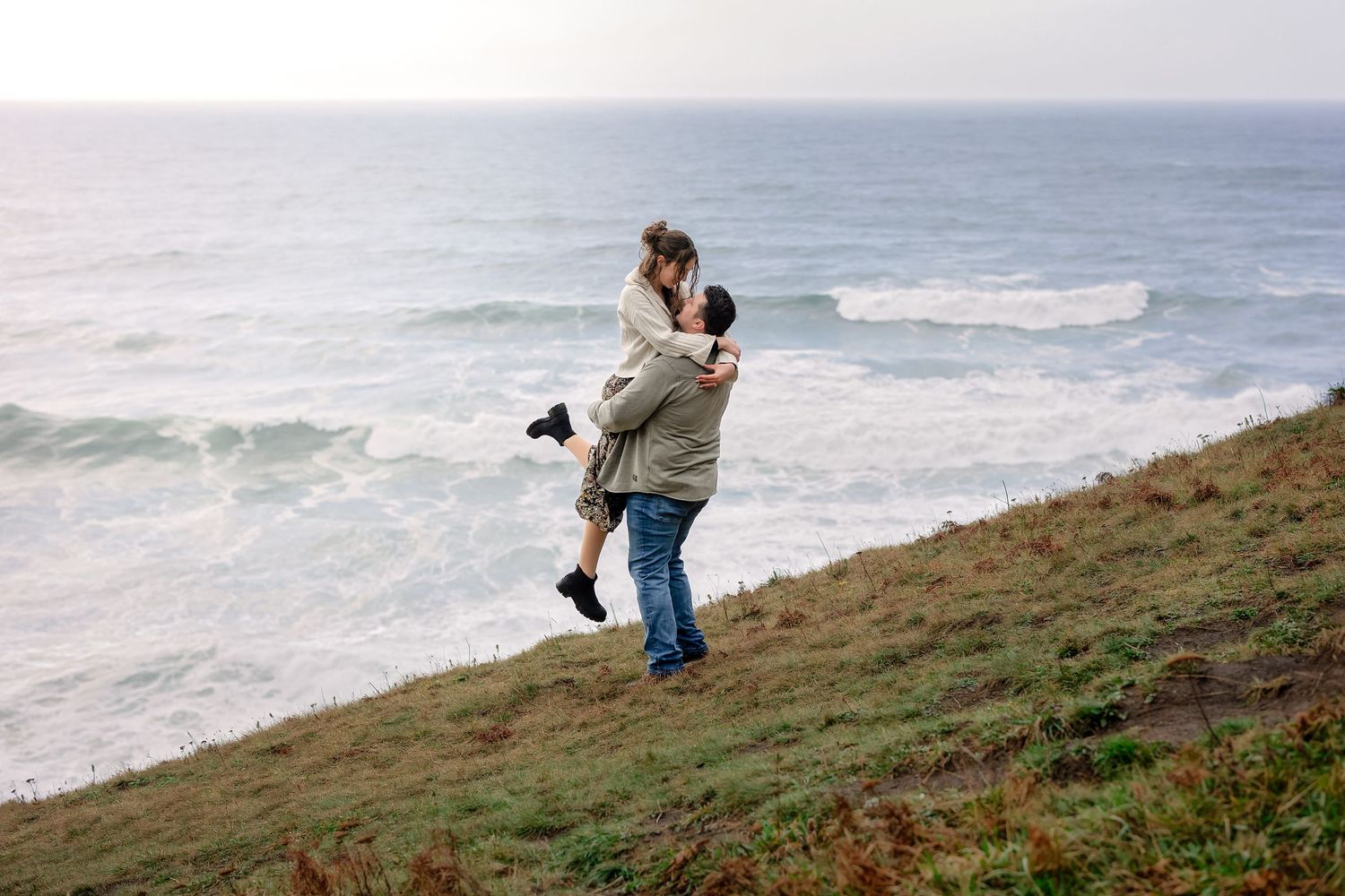 Couple shares romantic moment on coastal cliffside overlooking ocean waves during sunset.