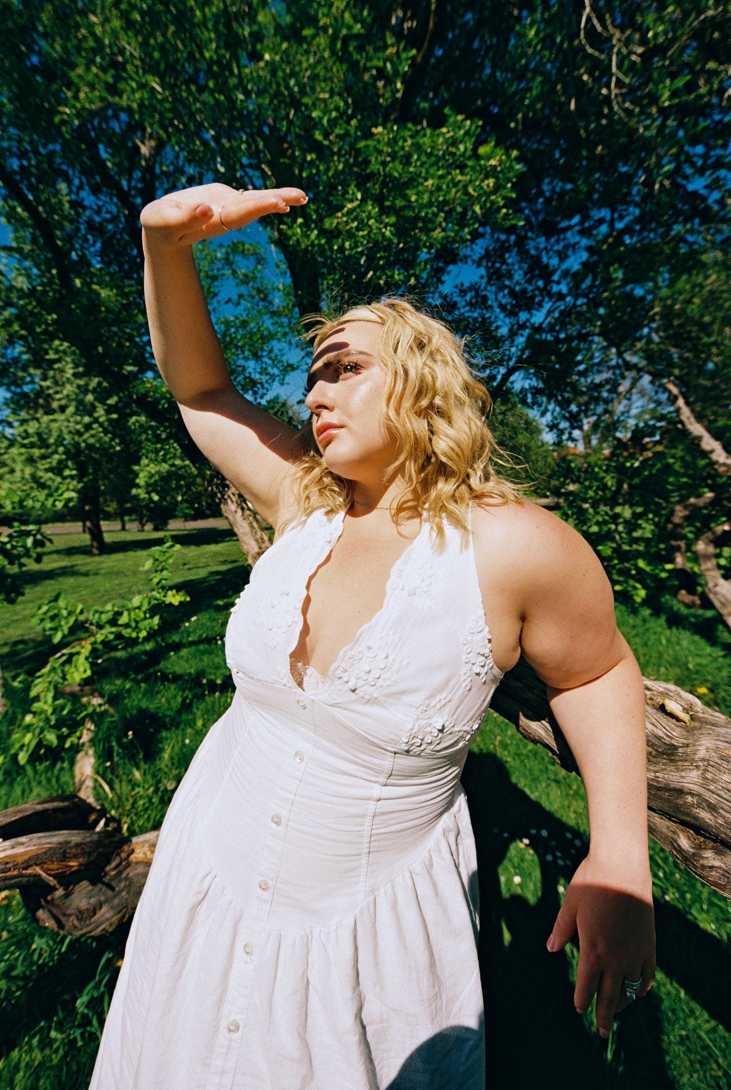 Woman in white sundress shielding eyes from sun, standing by rustic fence surrounded by lush green trees.