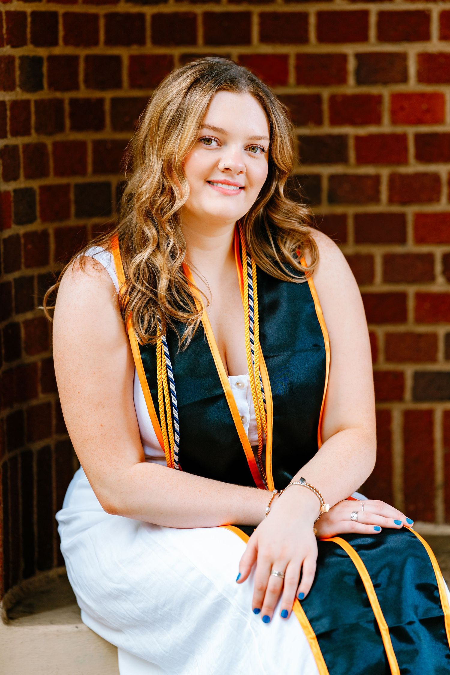 Young woman in graduation regalia with gold cords smiles while seated against a brick wall outdoors.