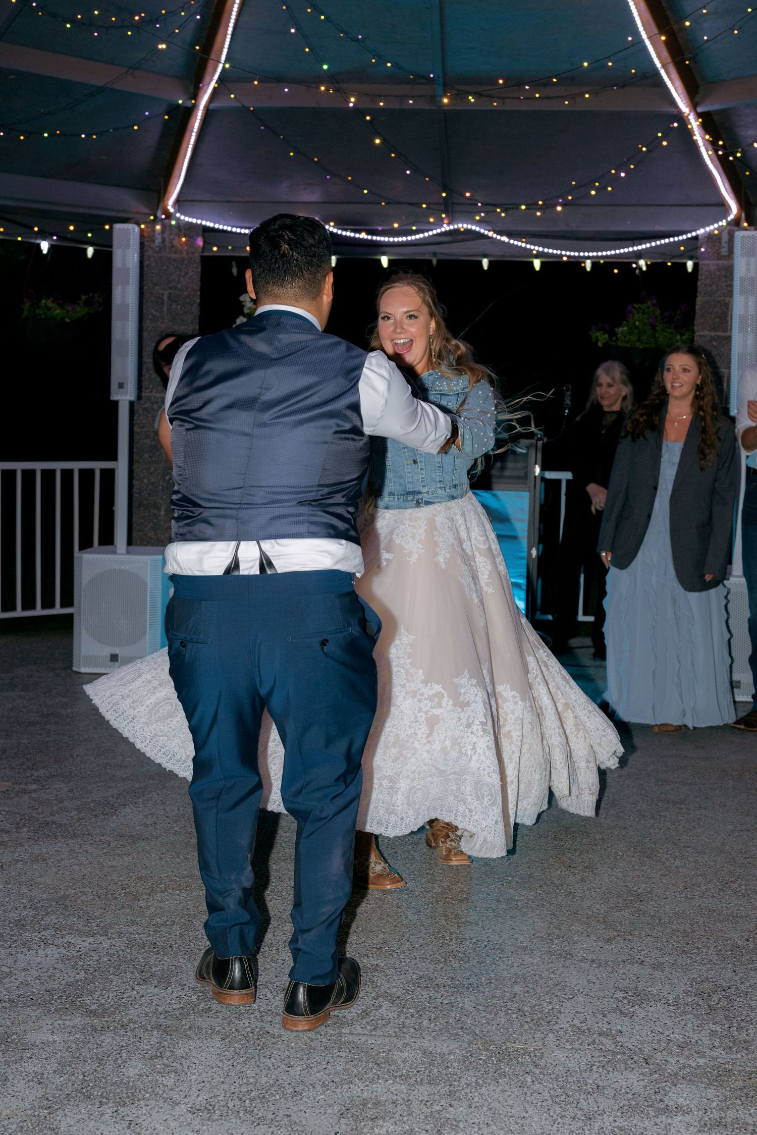 Bride in lace wedding dress and denim jacket shares first dance with groom under string lights.