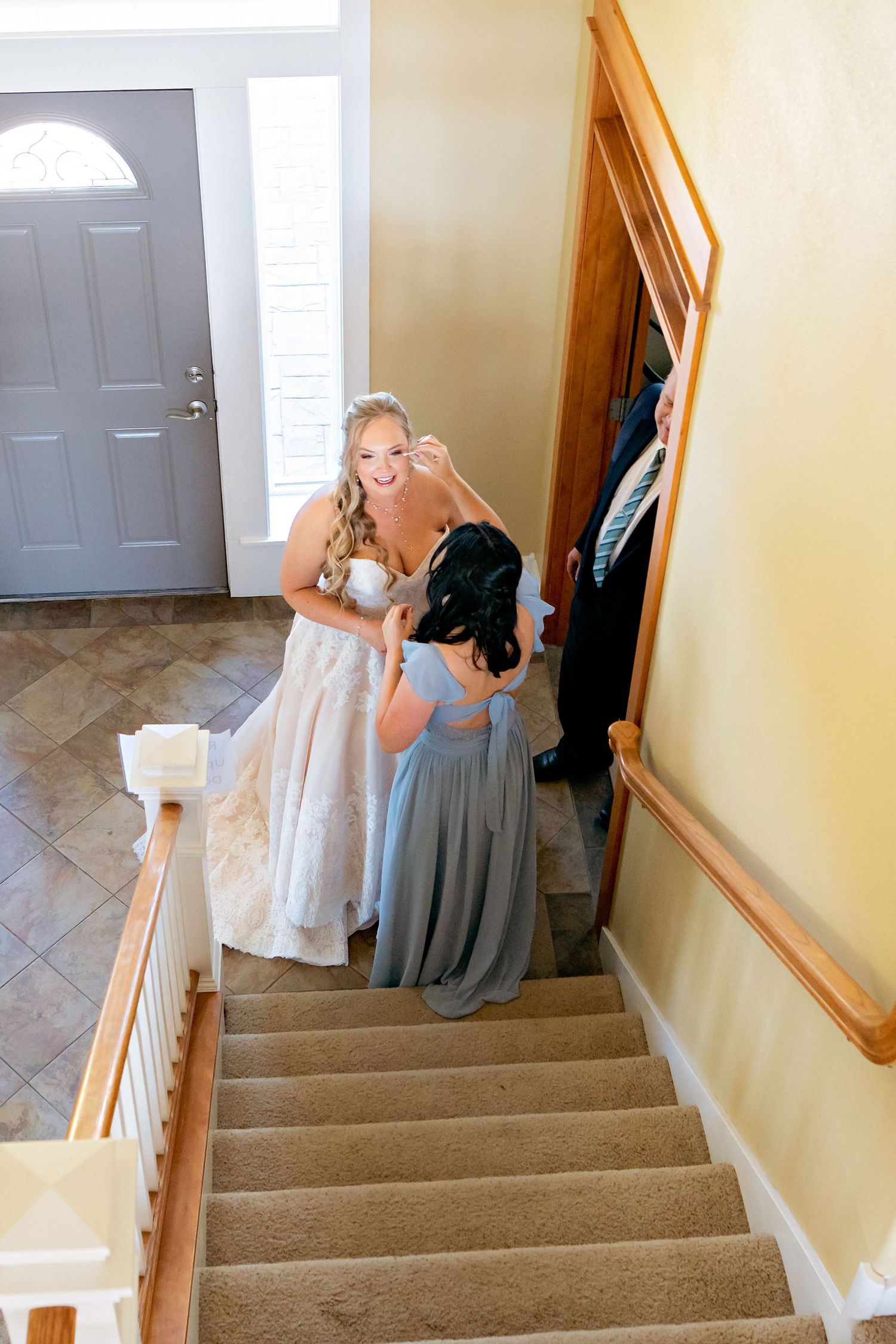 Smiling bride in lace wedding gown assisted by bridesmaid in blue dress on home staircase.