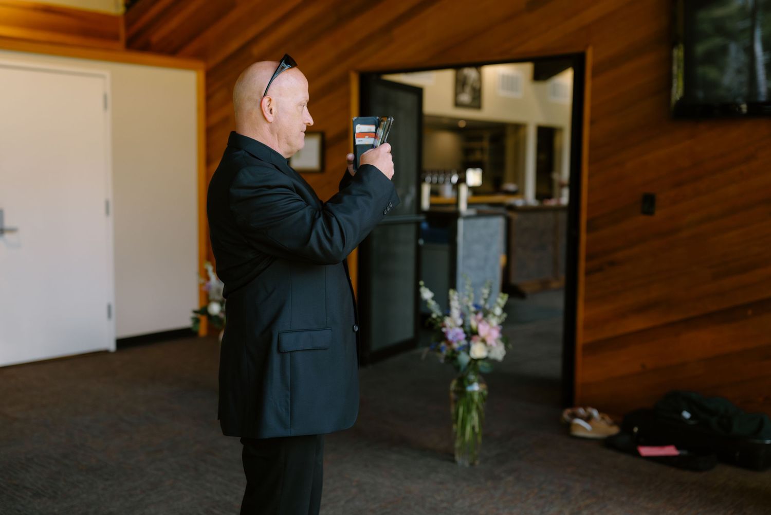 Man in black suit photographing floral arrangement in vase inside wooden venue during wedding event.