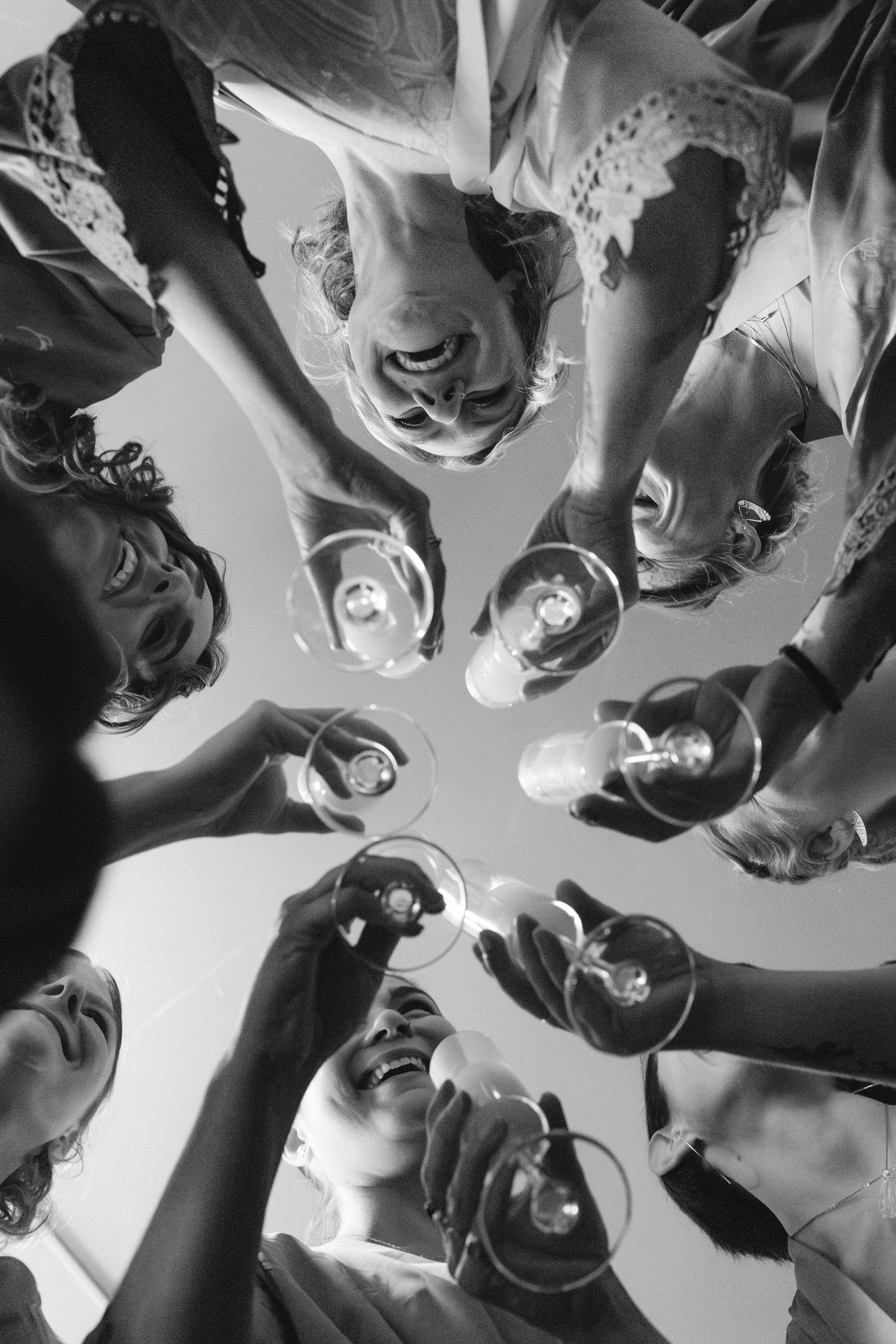 A black and white low angle view of people in a circle toasting with wine glasses against a bright background.