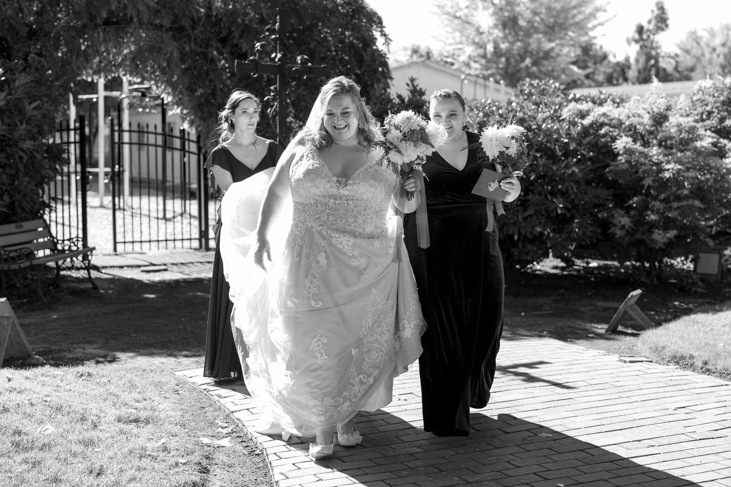 A bride in a flowing white dress walks with attendants in black dresses along a garden pathway near a decorative gate.