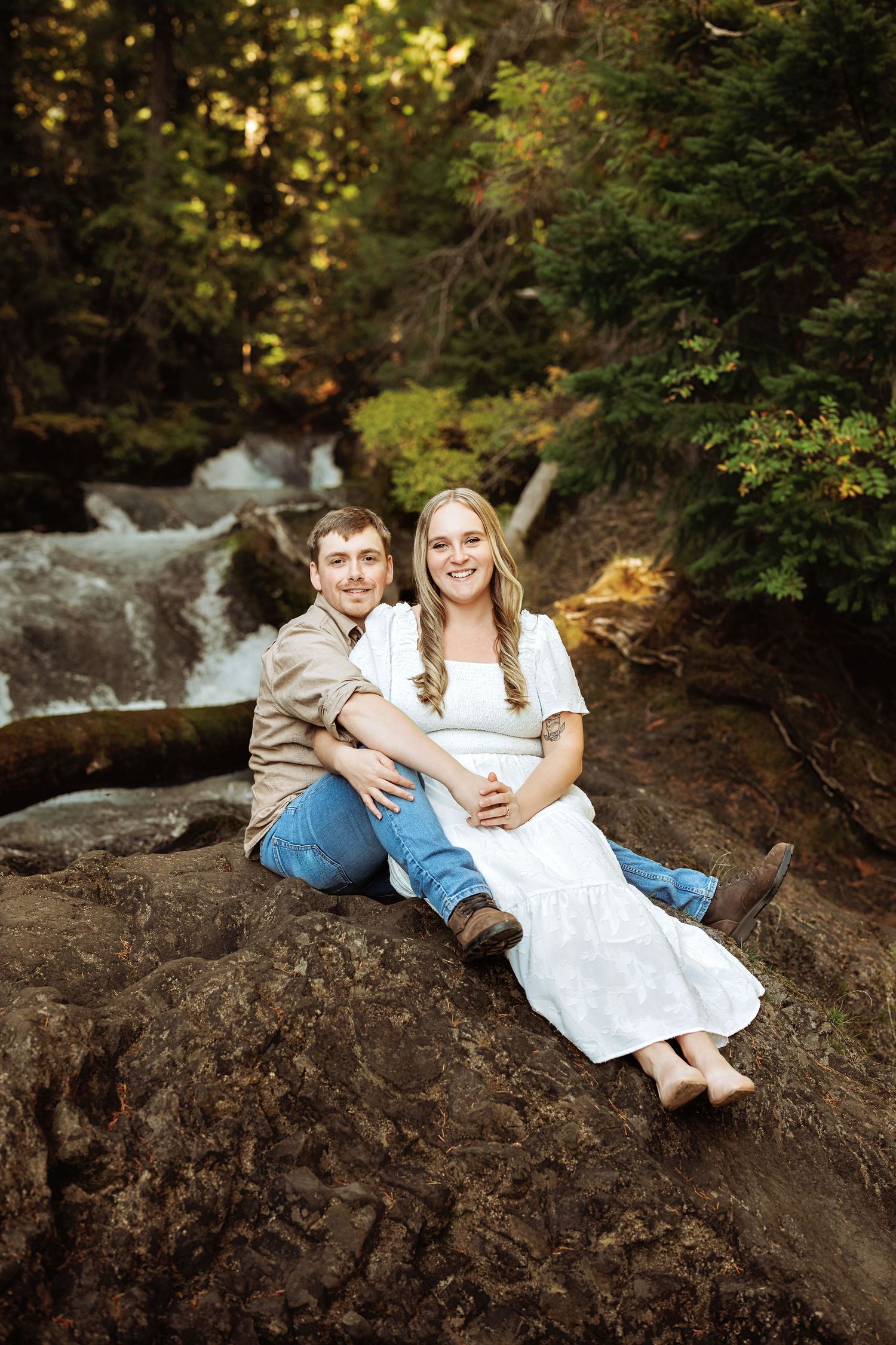 Couple relaxing on rocky outcrop by rushing stream in wooded setting during outdoor photoshoot.