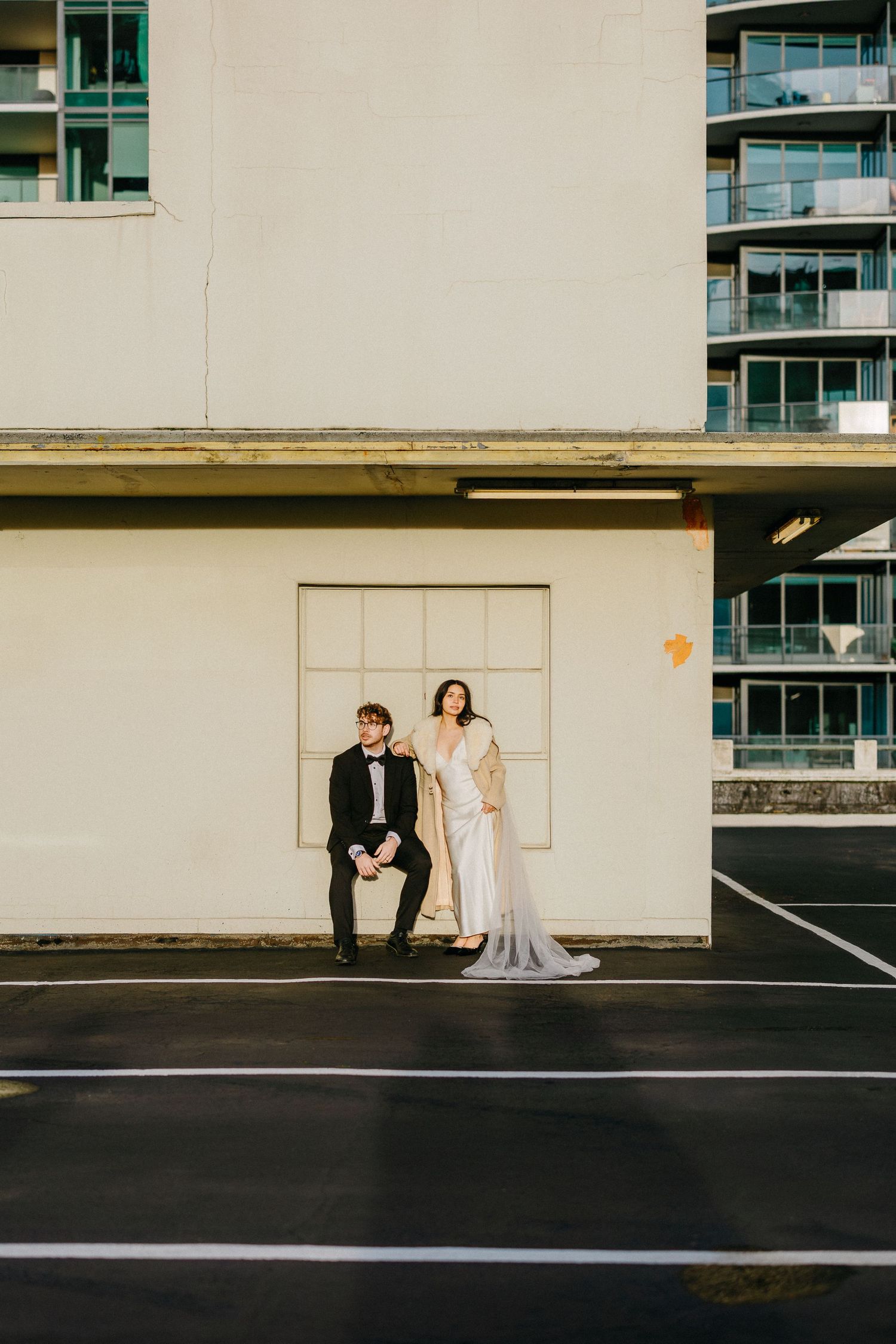 Artistic wedding couple poses against white wall in minimalist urban setting with modern architecture.