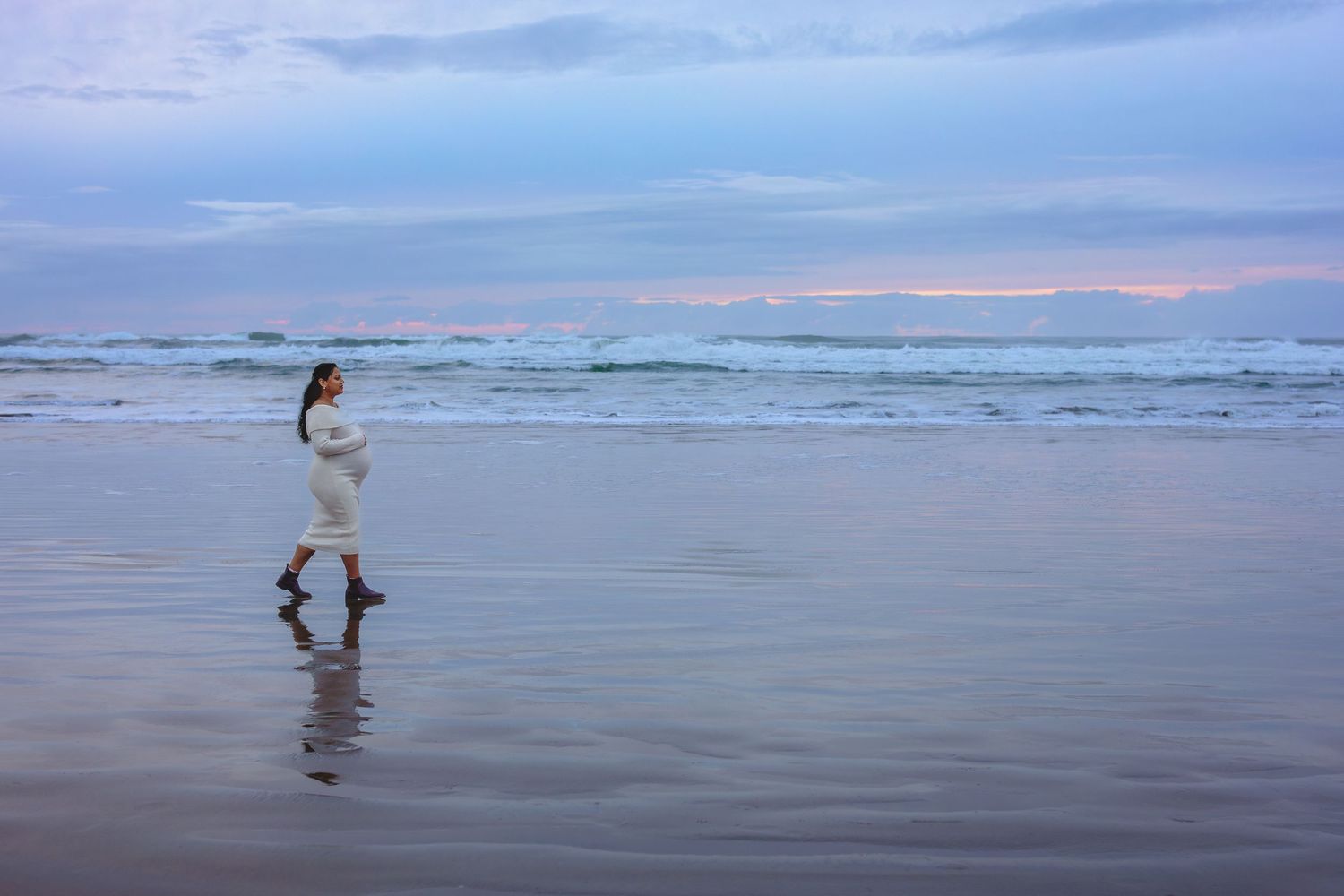 A solitary figure walks along a misty beach at dusk, creating reflections in the wet sand as waves roll in from the ocean.
