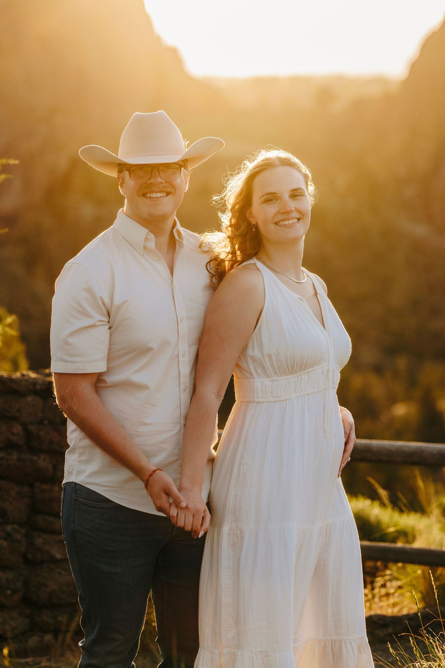 A couple poses together at sunset, with her in a white dress and him in a cowboy hat against rocky cliffs.