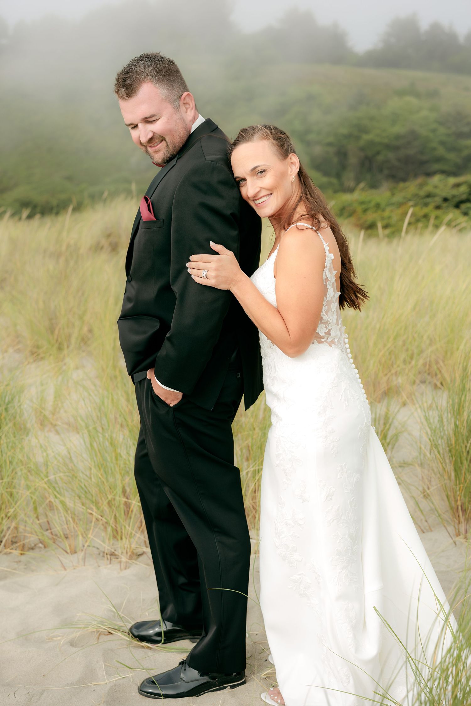 Couple shares playful moment on sandy beach with foggy coastal hills behind them.