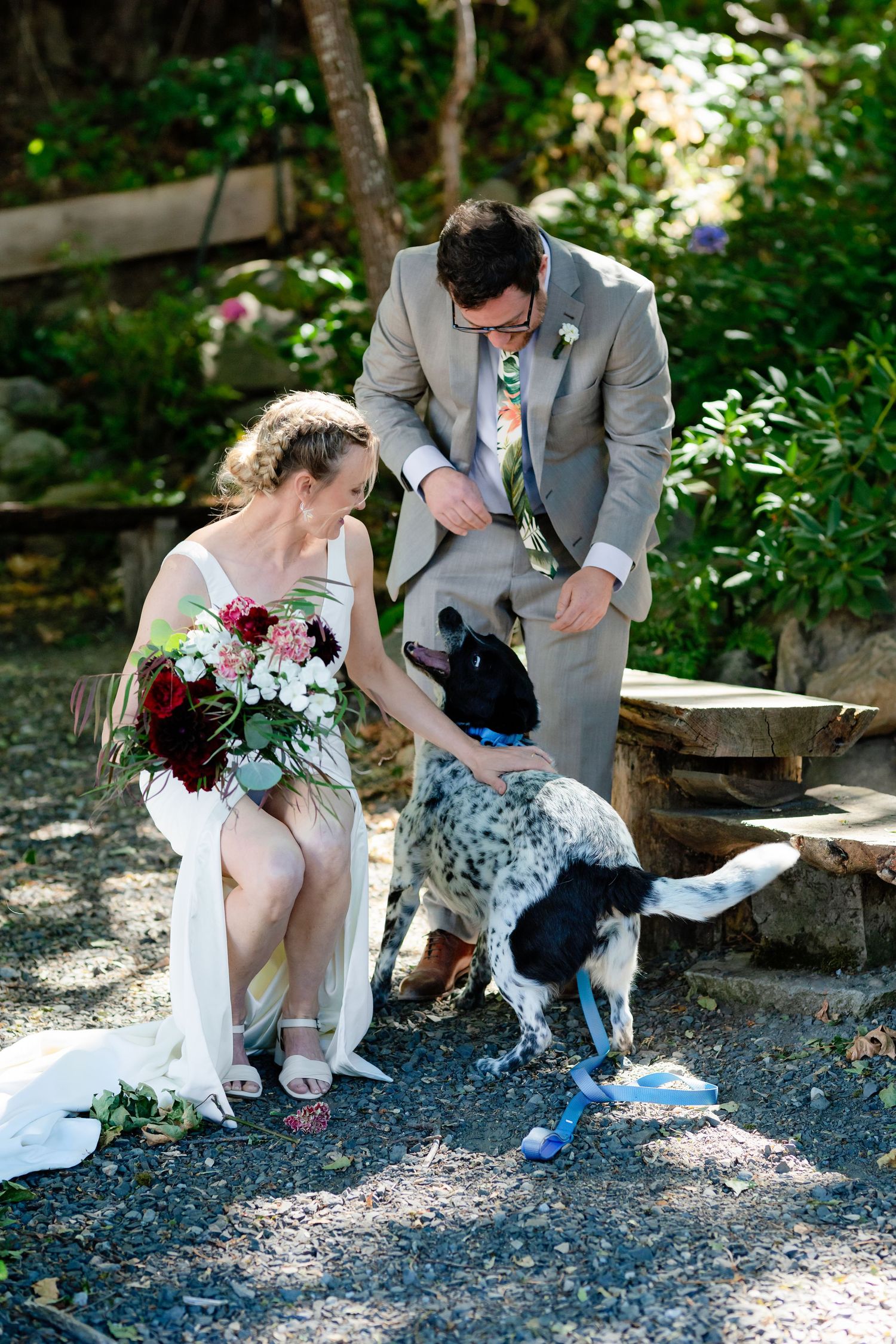Bride and groom crouch down to pet their black and white dog during outdoor wedding photos.