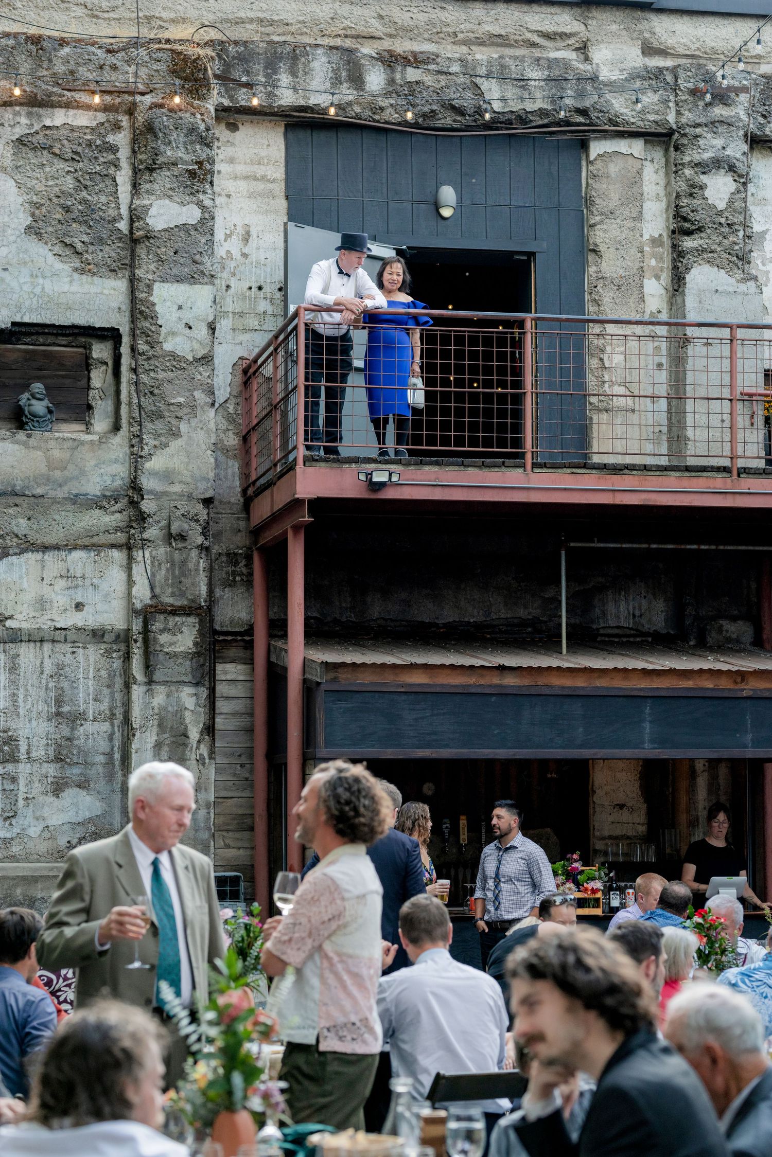 Wedding guests mingle at outdoor reception while couple overlooks from industrial balcony of rustic stone venue.