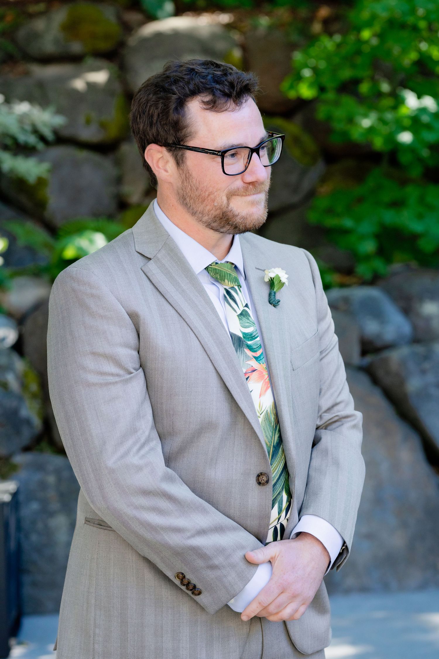 Groom in gray suit with tropical floral tie and boutonniere stands attentively at outdoor ceremony.