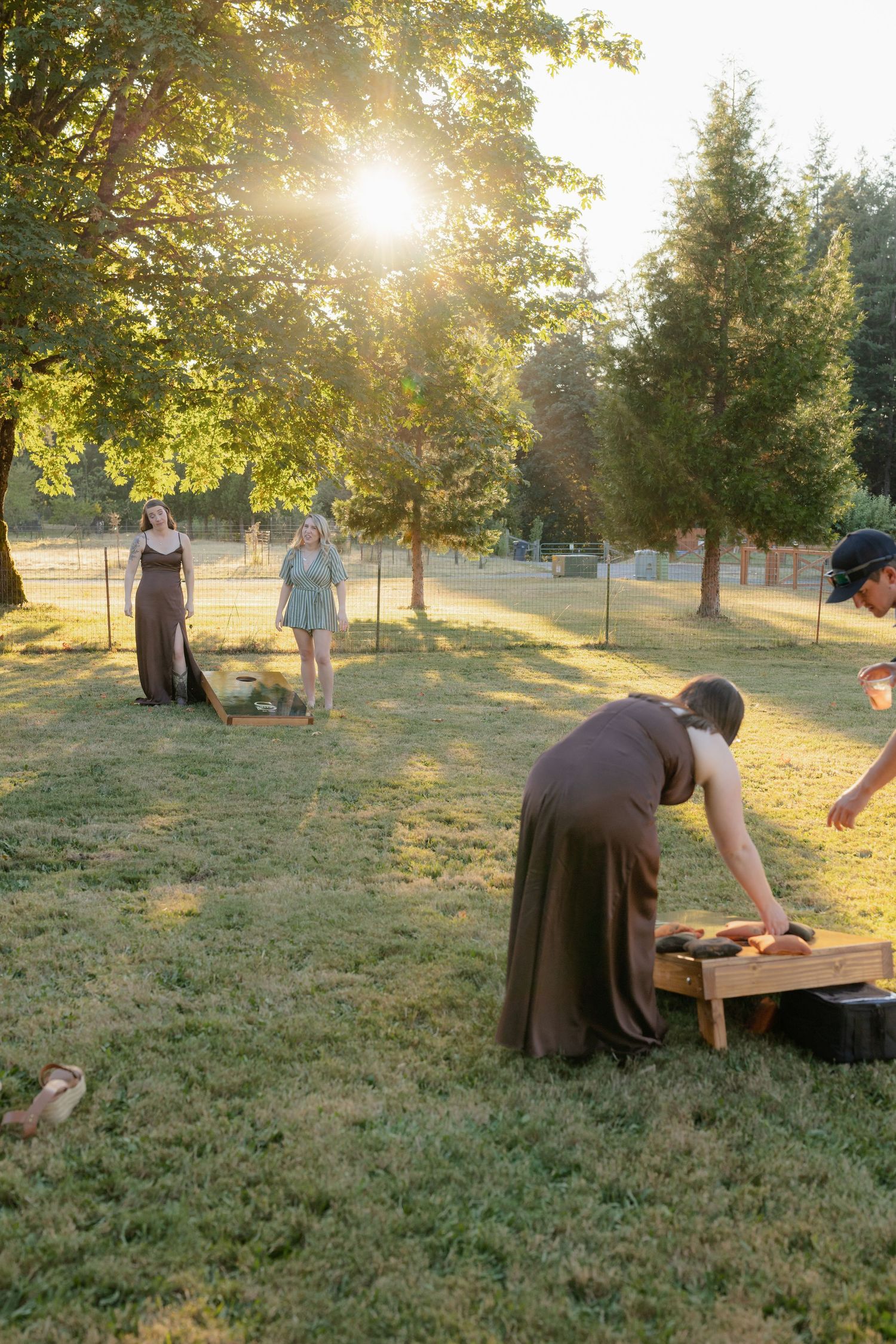 Wedding guests playing cornhole lawn game at golden hour, with bridesmaids in brown dresses enjoying outdoor reception activities.