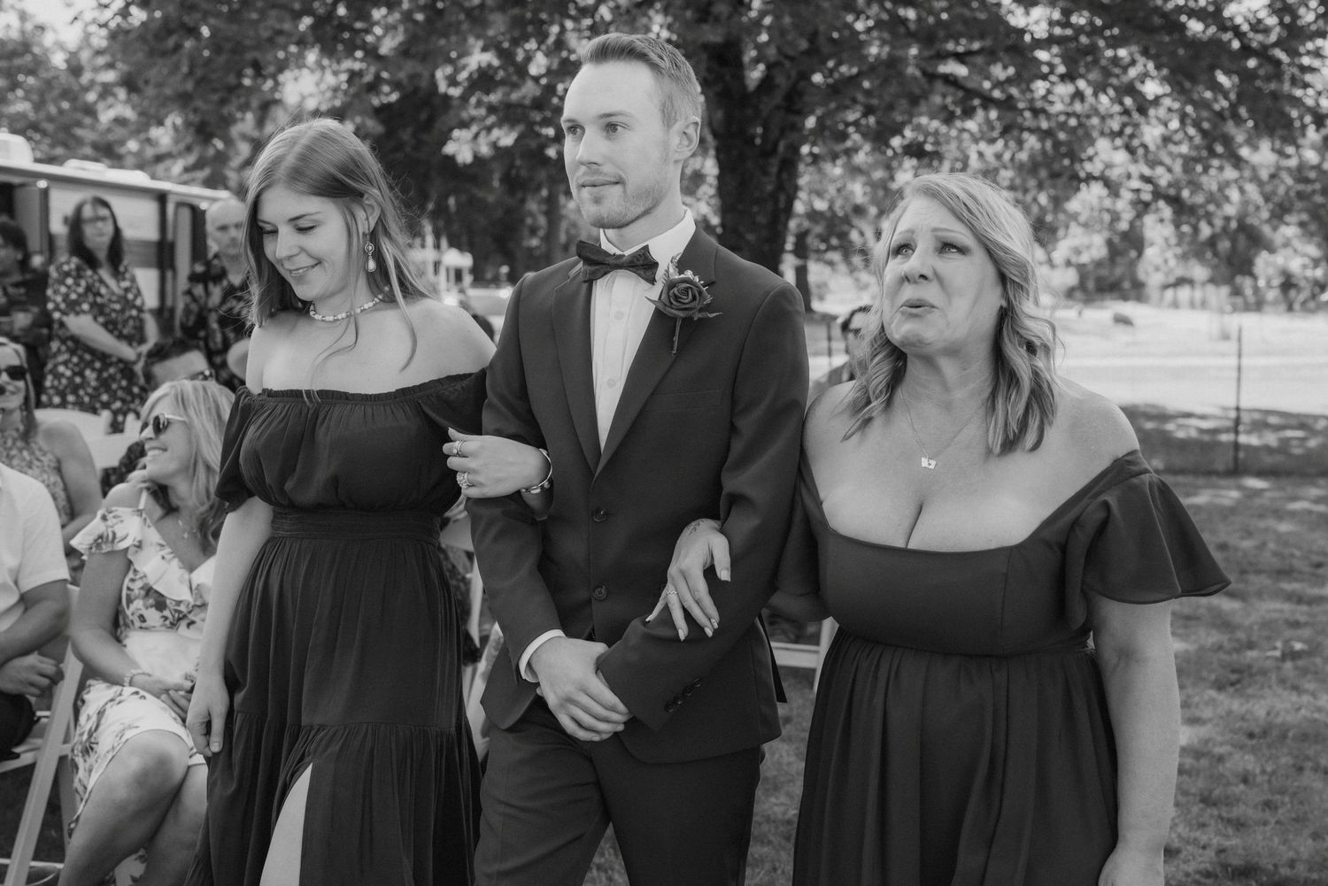 A groom in a tuxedo walks down the aisle escorted by two women in black off-shoulder dresses at a wedding.