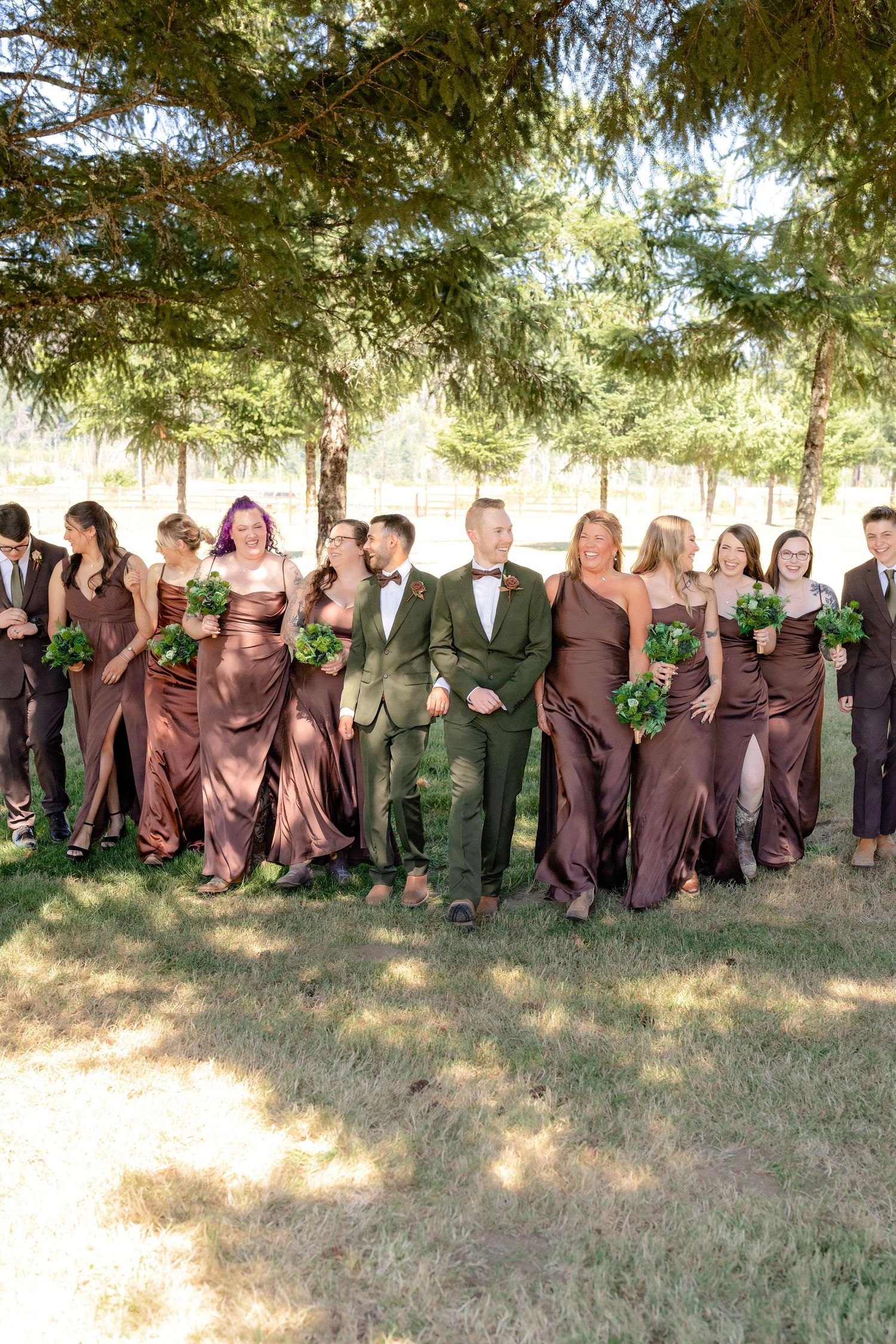 Two grooms in green suits walk with wedding party in brown satin dresses under pine trees.