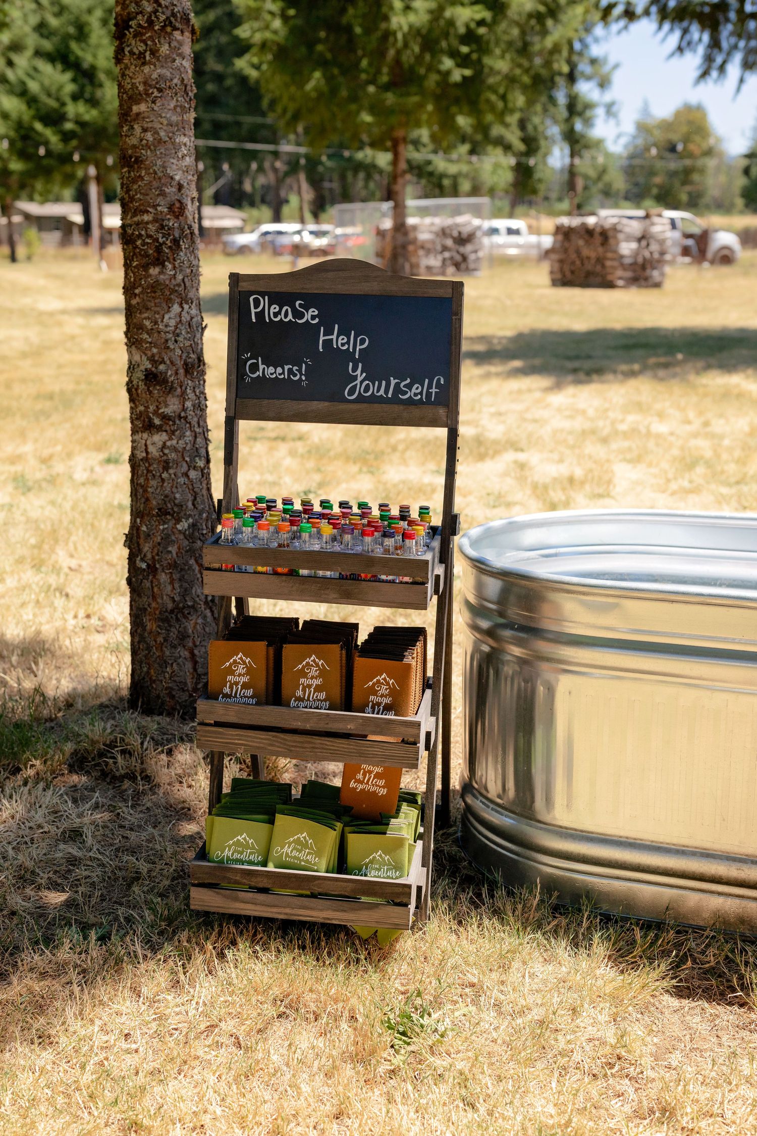 Rustic wooden drink station with colorful beverages and a chalkboard sign reading Please Help Yourself next to a metal tub outdoors.