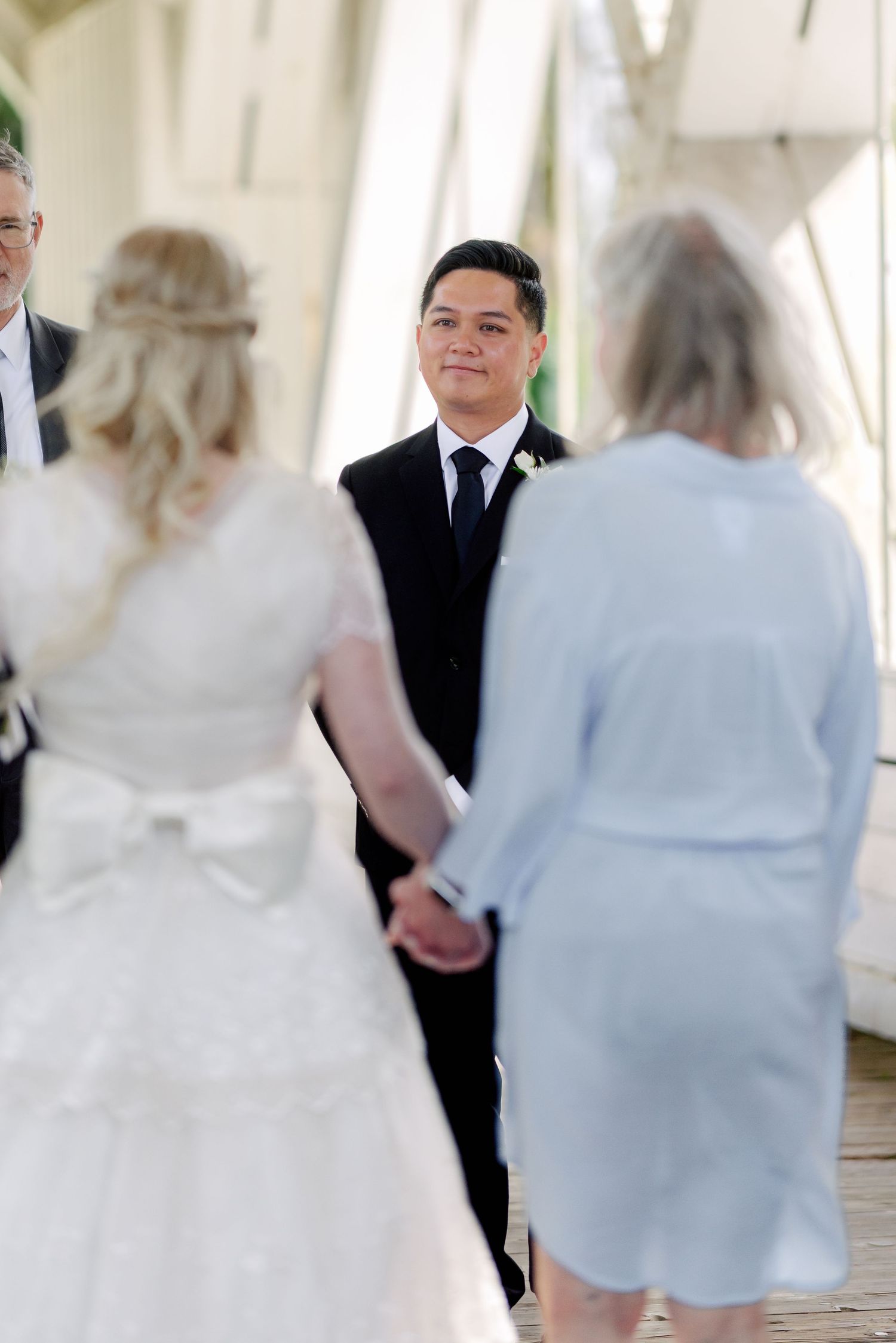 Groom in black suit smiles at bride during outdoor wedding ceremony while holding hands.