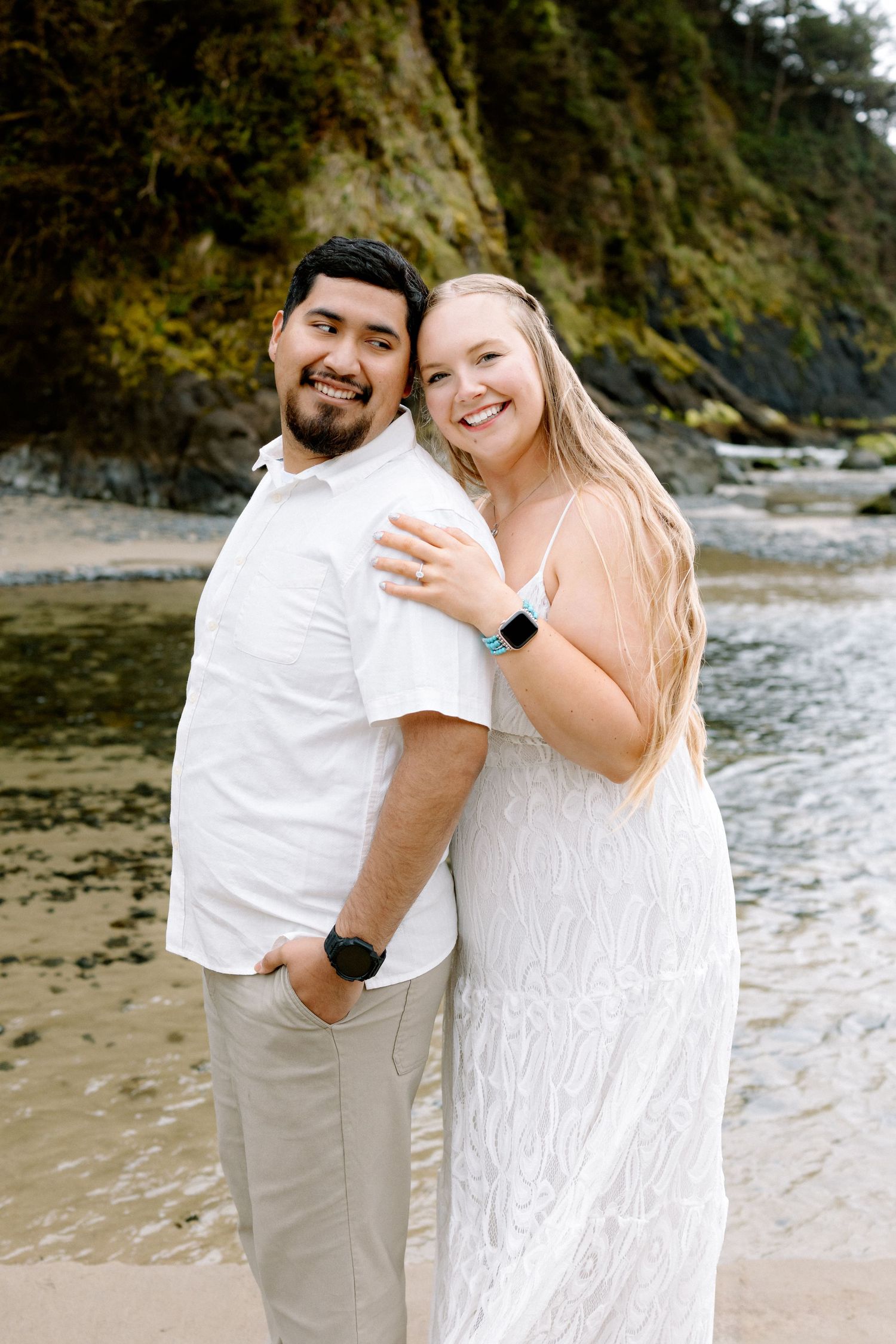 Happy couple posing together on beach, woman in white lace dress hugging man in white shirt.