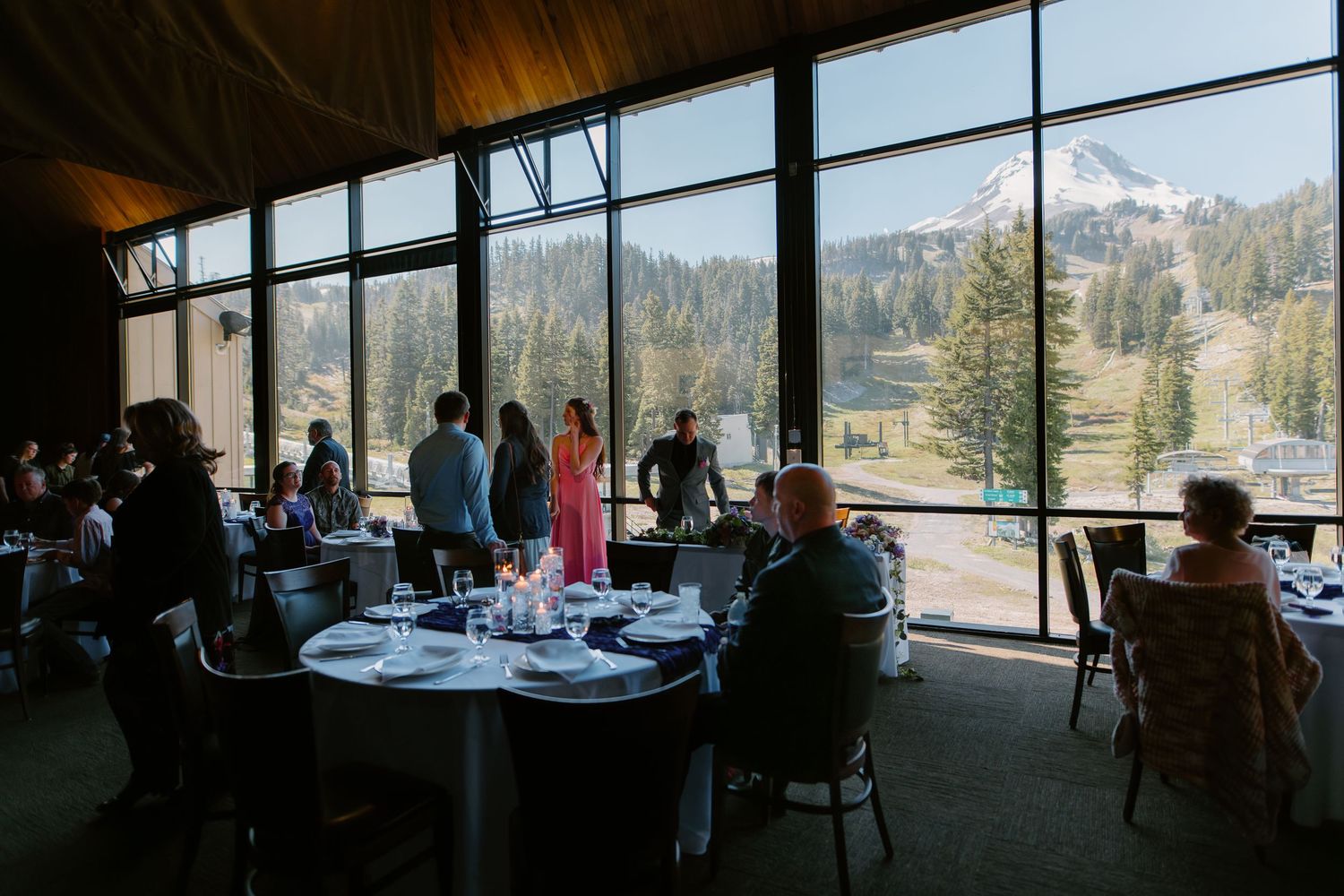 Elegant mountain lodge dining room with floor-to-ceiling windows overlooking snowy peaks and guests gathered for an event.