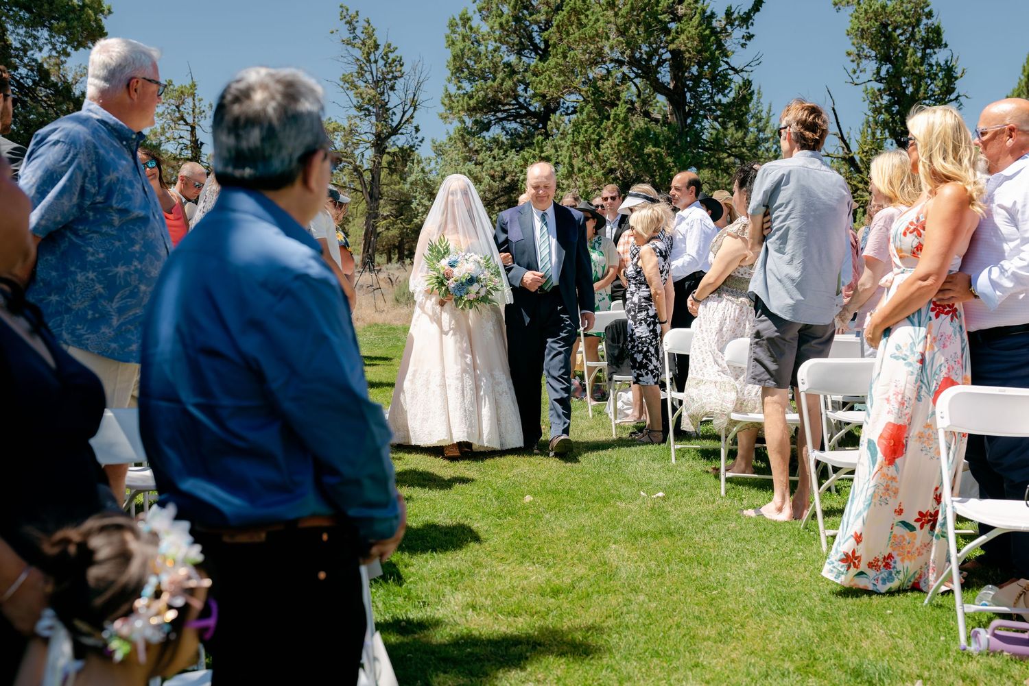 Bride walks down outdoor aisle with escort at sunny wedding ceremony surrounded by seated guests.