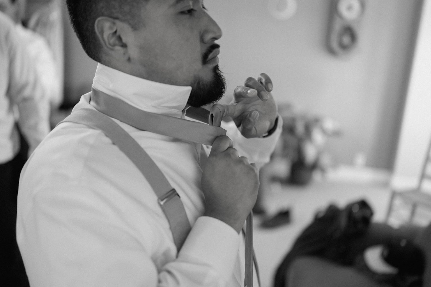 Groom in white shirt and suspenders adjusting his bow tie in black and white photo.