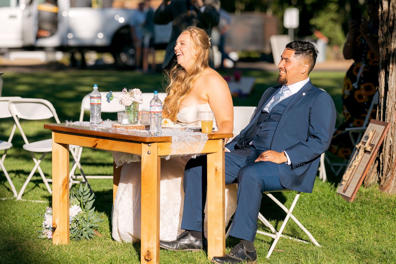 Bride and groom sit together at sweetheart table laughing during outdoor wedding reception.