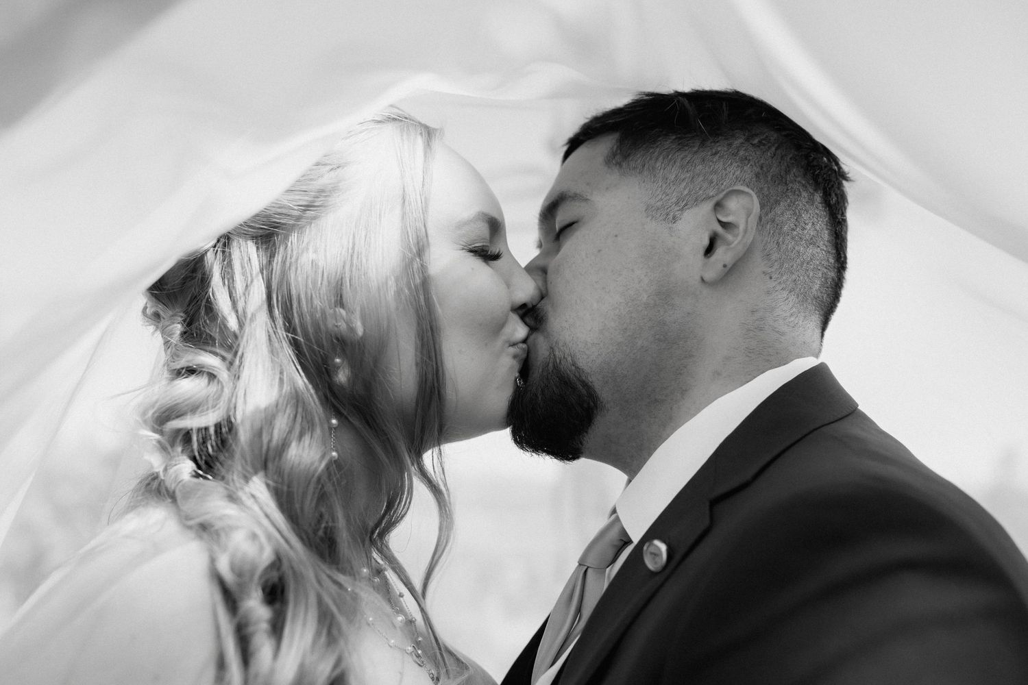 Black and white romantic photo of a bride and groom sharing a kiss under a flowing wedding veil.