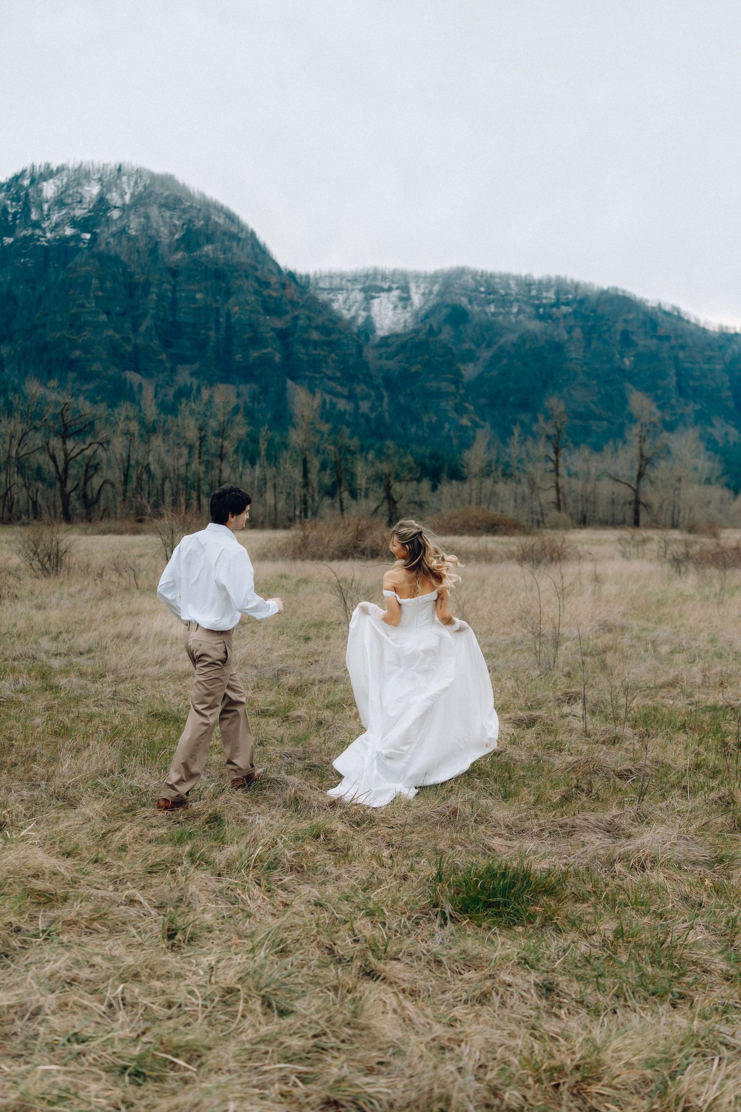 A couple in wedding attire runs playfully through a field with mountains in the background.