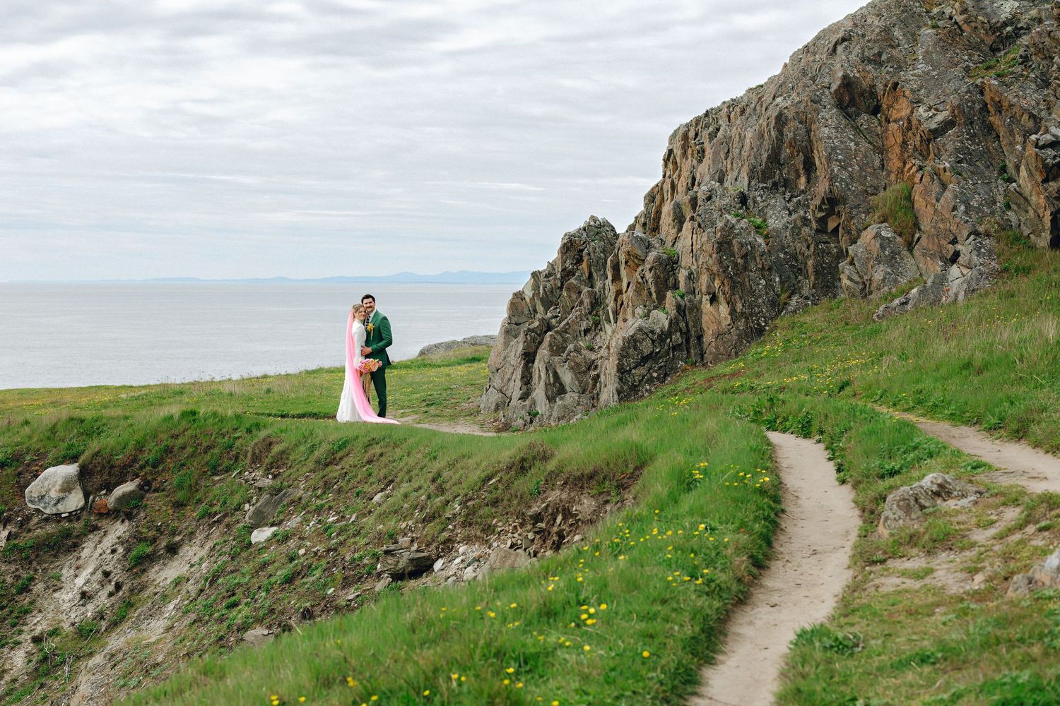 Couple embraces on scenic coastal cliff path with ocean views and green hillside landscape.
