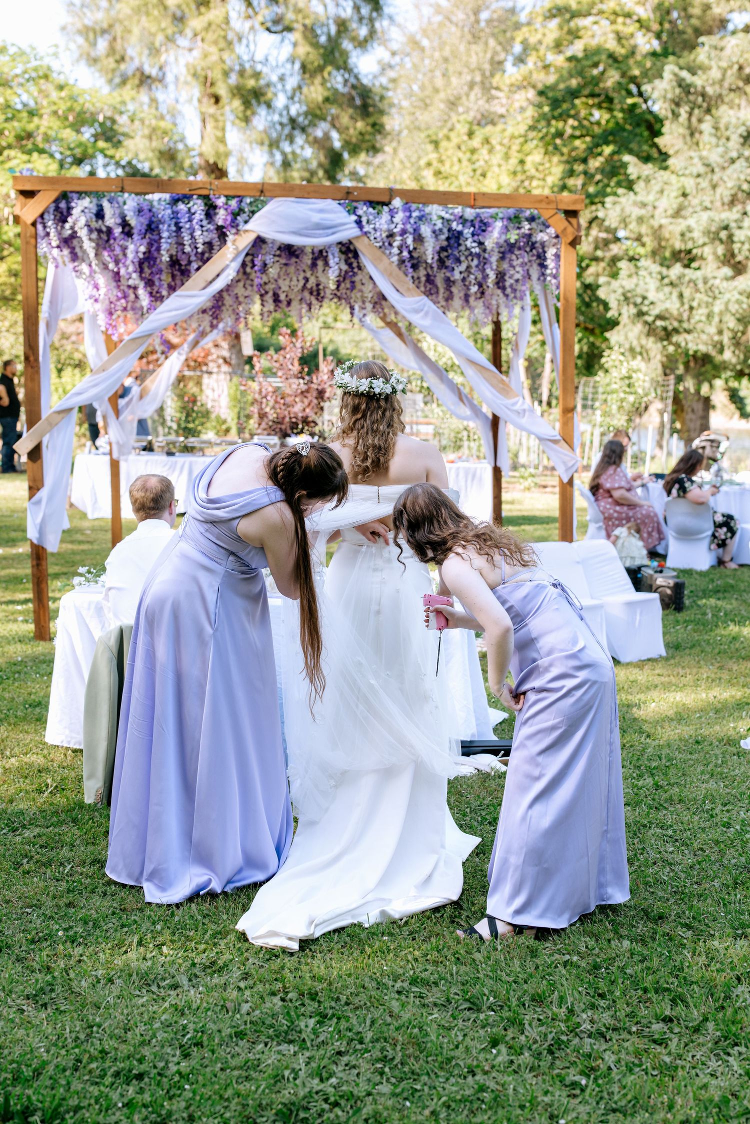 Bridesmaids in lavender dresses adjust the brides gown beneath a wooden arch adorned with purple wisteria.