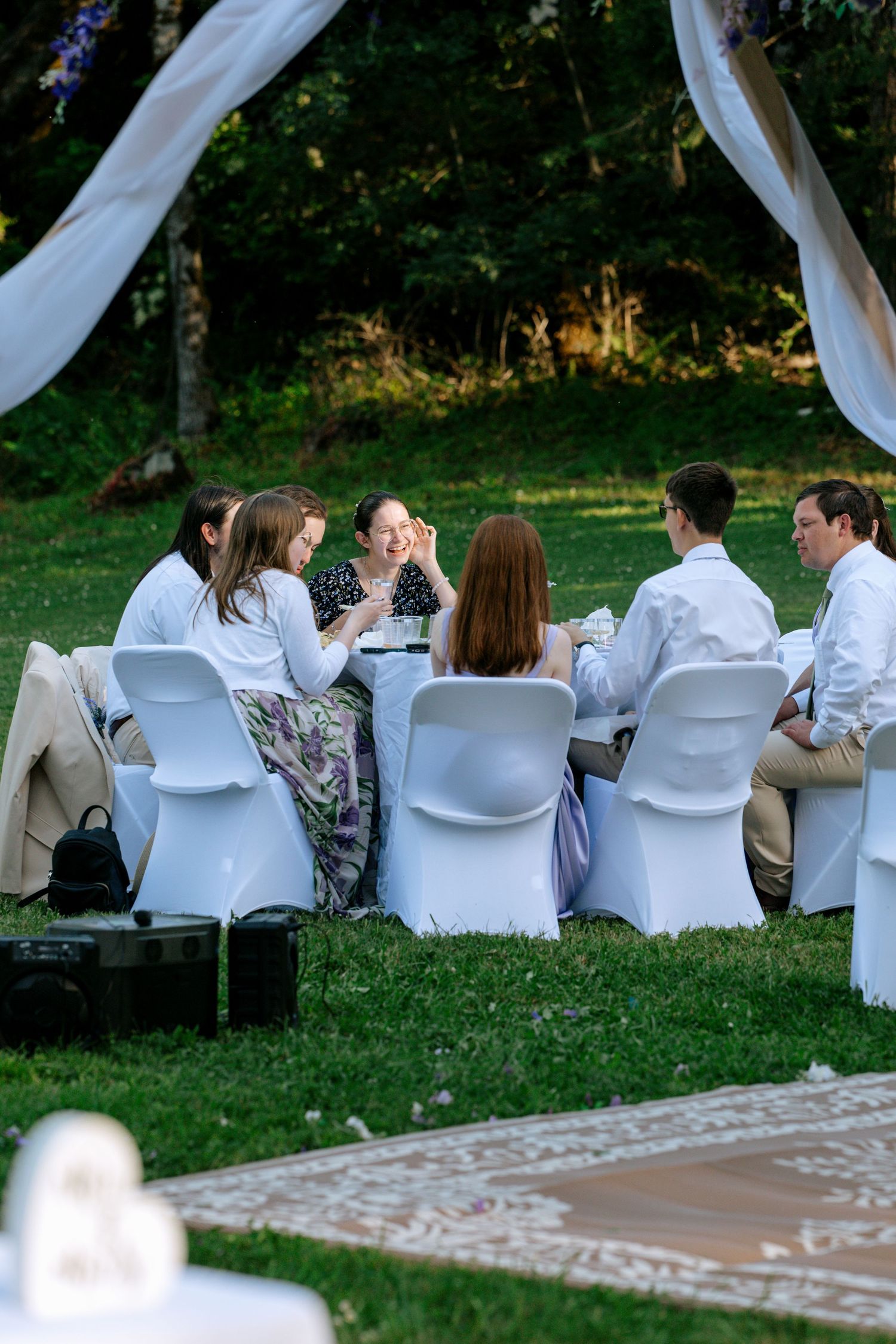 Wedding guests laugh and socialize at outdoor reception table under white draped canopy at garden wedding.