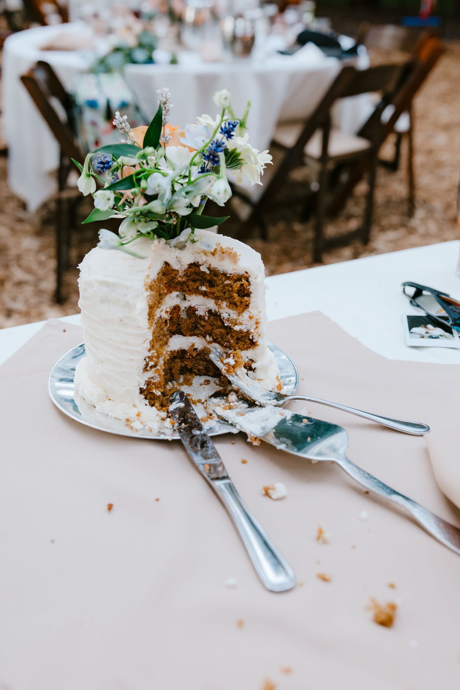 Partially sliced three-layer carrot wedding cake with white frosting and floral topper on silver stand.