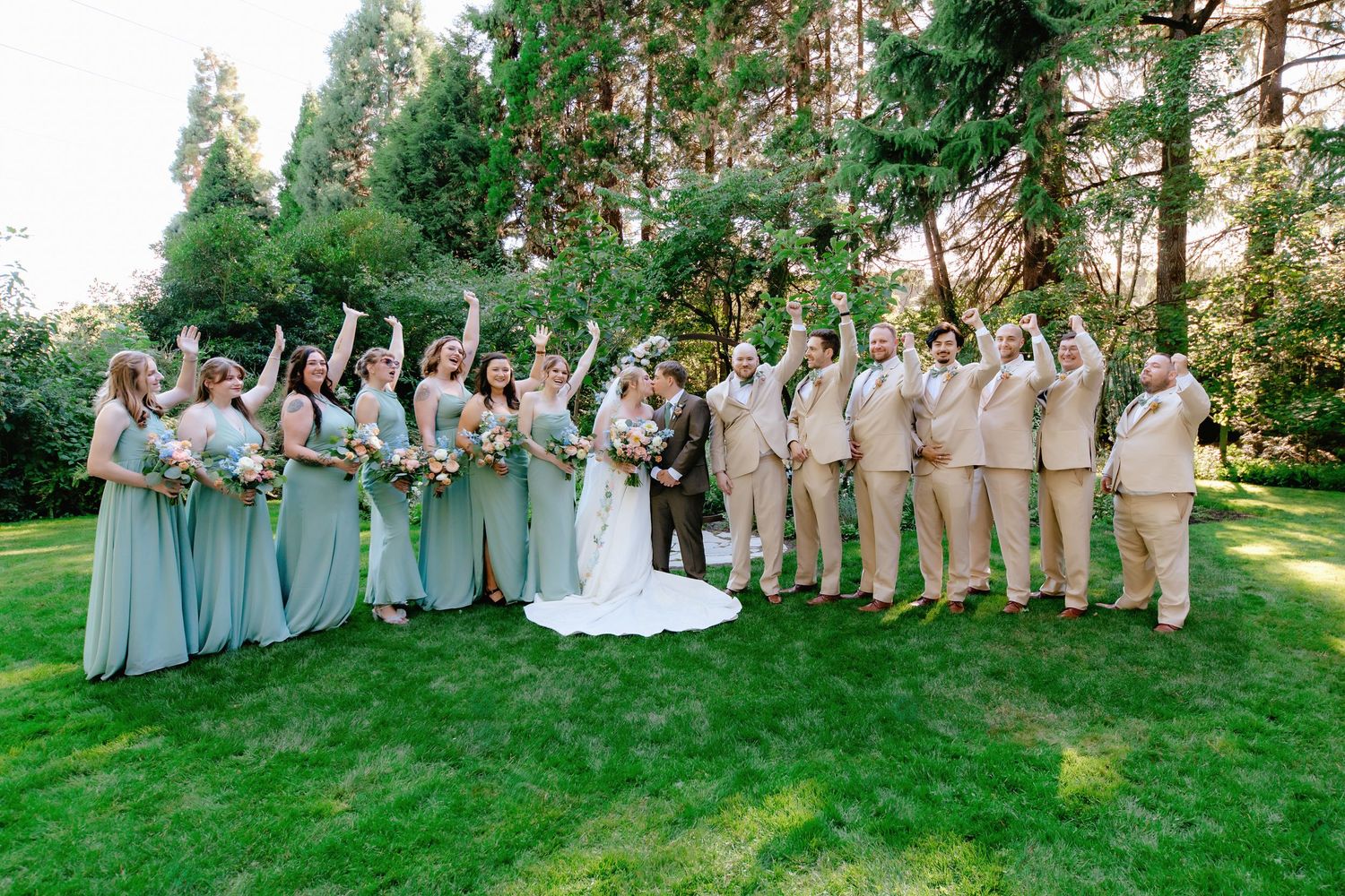Large wedding party in sage green and tan attire celebrates on green lawn surrounded by tall trees.
