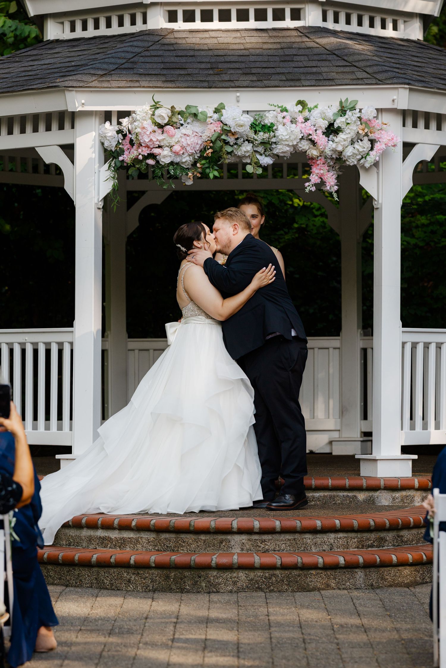 Bride and groom share first kiss under floral-adorned white gazebo at outdoor wedding ceremony.