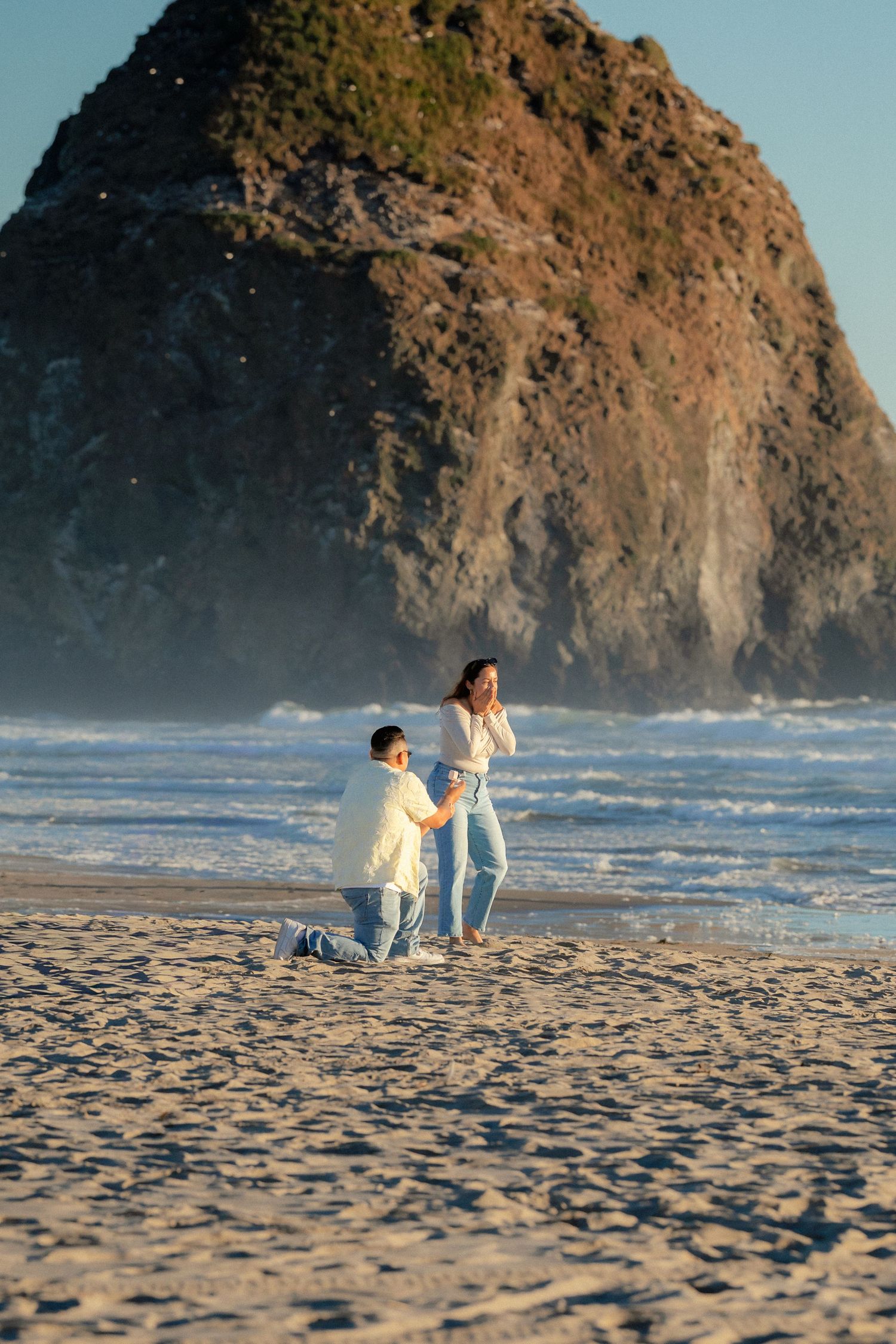 Man proposing to woman on sandy beach with dramatic rock formation and ocean waves at golden hour.