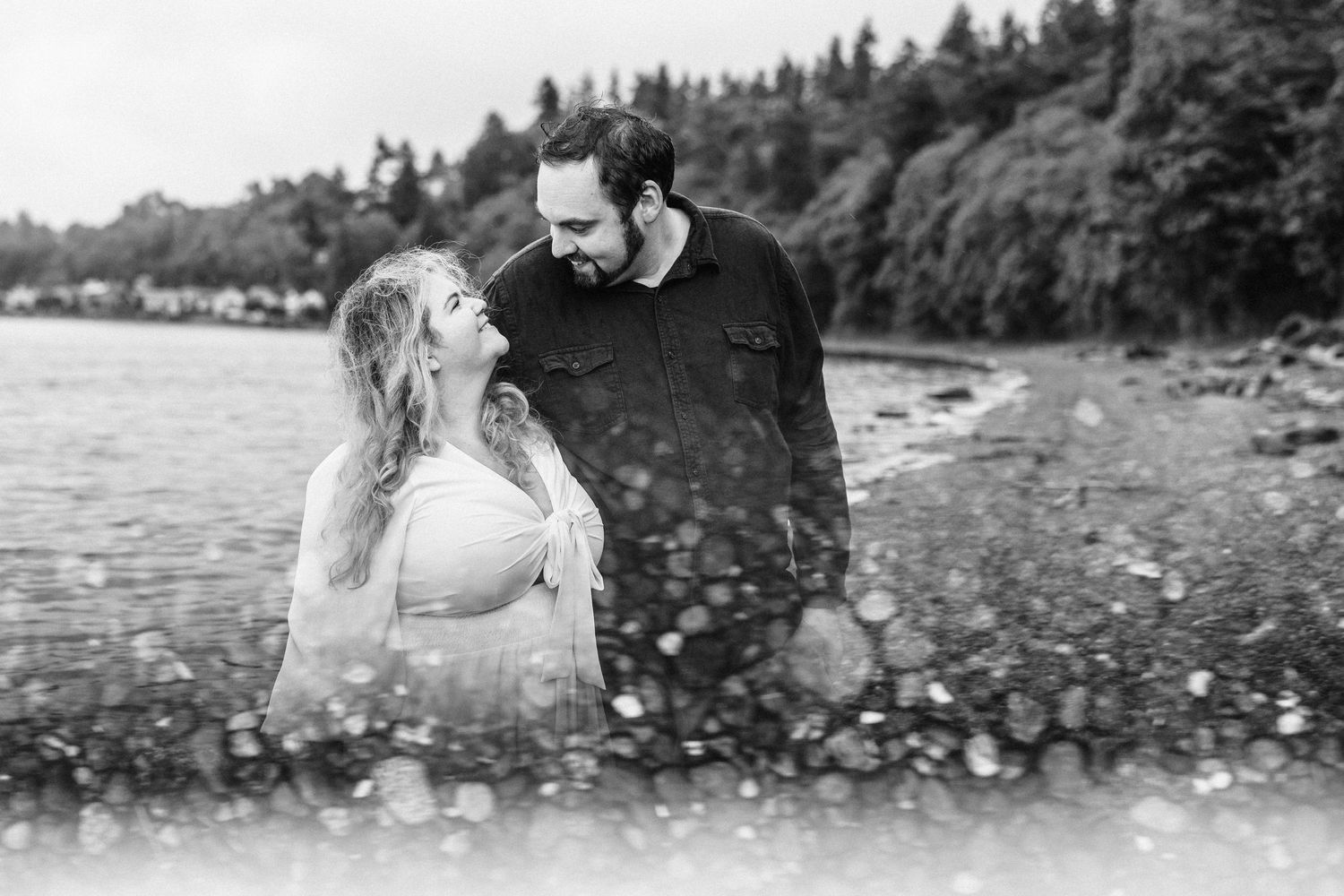 A romantic couple embraces on a rocky beach with evergreen trees and mountains in the background.