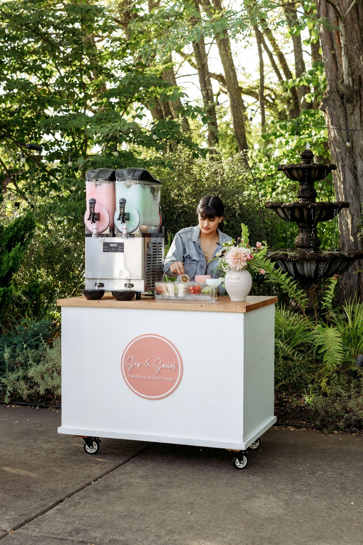 Mobile white bar cart with pink signage set up outdoors against a lush green garden backdrop.